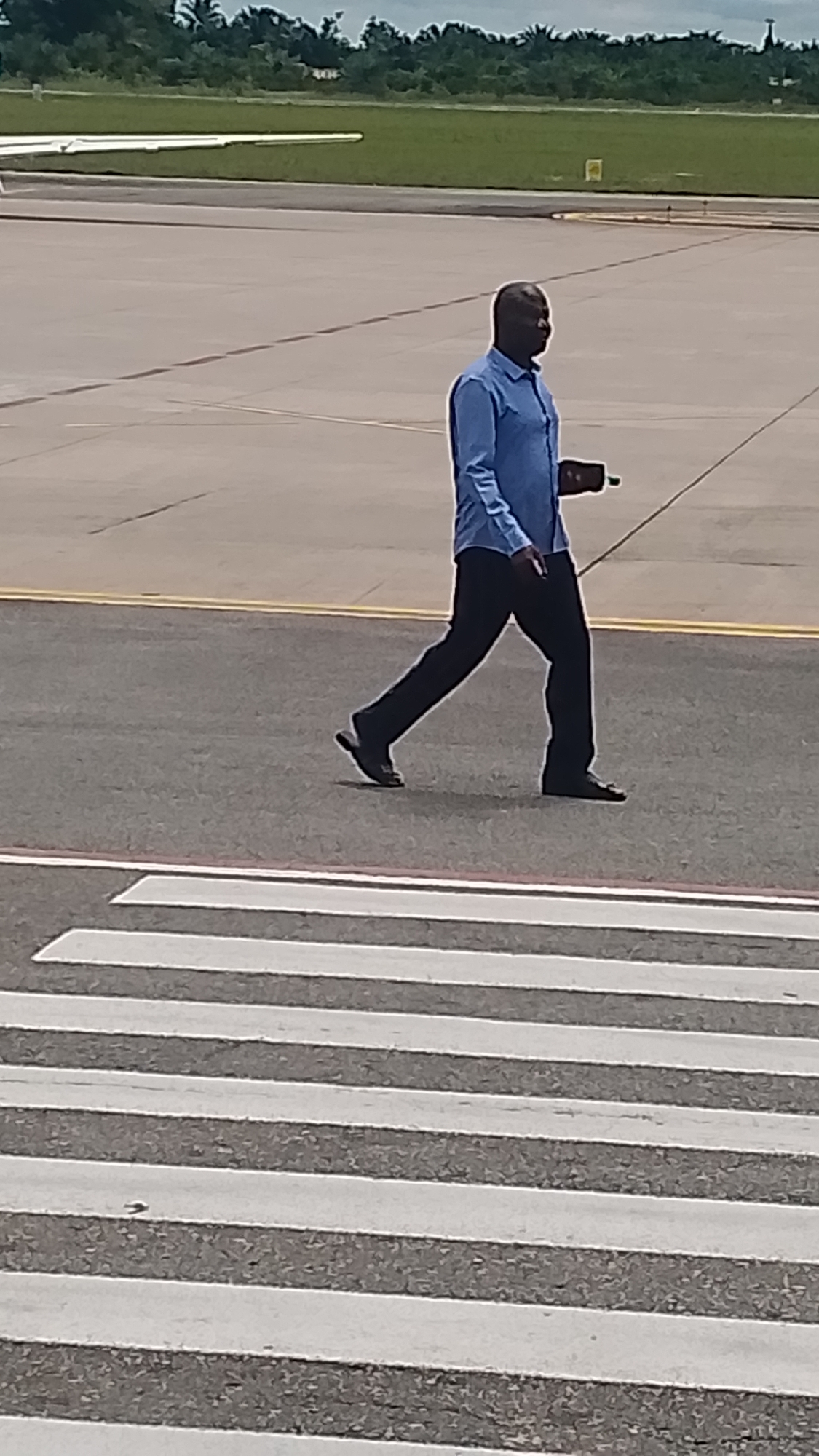 A medium-shot image shows a dark-skinned man walking across a tarmac at what appears to be an airport in Orerokpe, Nigeria. He is wearing a blue collared shirt and dark pants, and is carrying a small, dark object in his right hand. He is walking from right to left in the frame, his gait suggesting he is moving with purpose. The tarmac is grey and marked with yellow lines. In the foreground, a white pedestrian crossing is visible. In the background, a portion of an airplane wing and lush green trees can be seen. The sky is overcast, suggesting it is likely daytime, possibly late morning or early afternoon, with diffused sunlight. The overall scene conveys a sense of movement and activity at an airport. There is no visible text in the image.