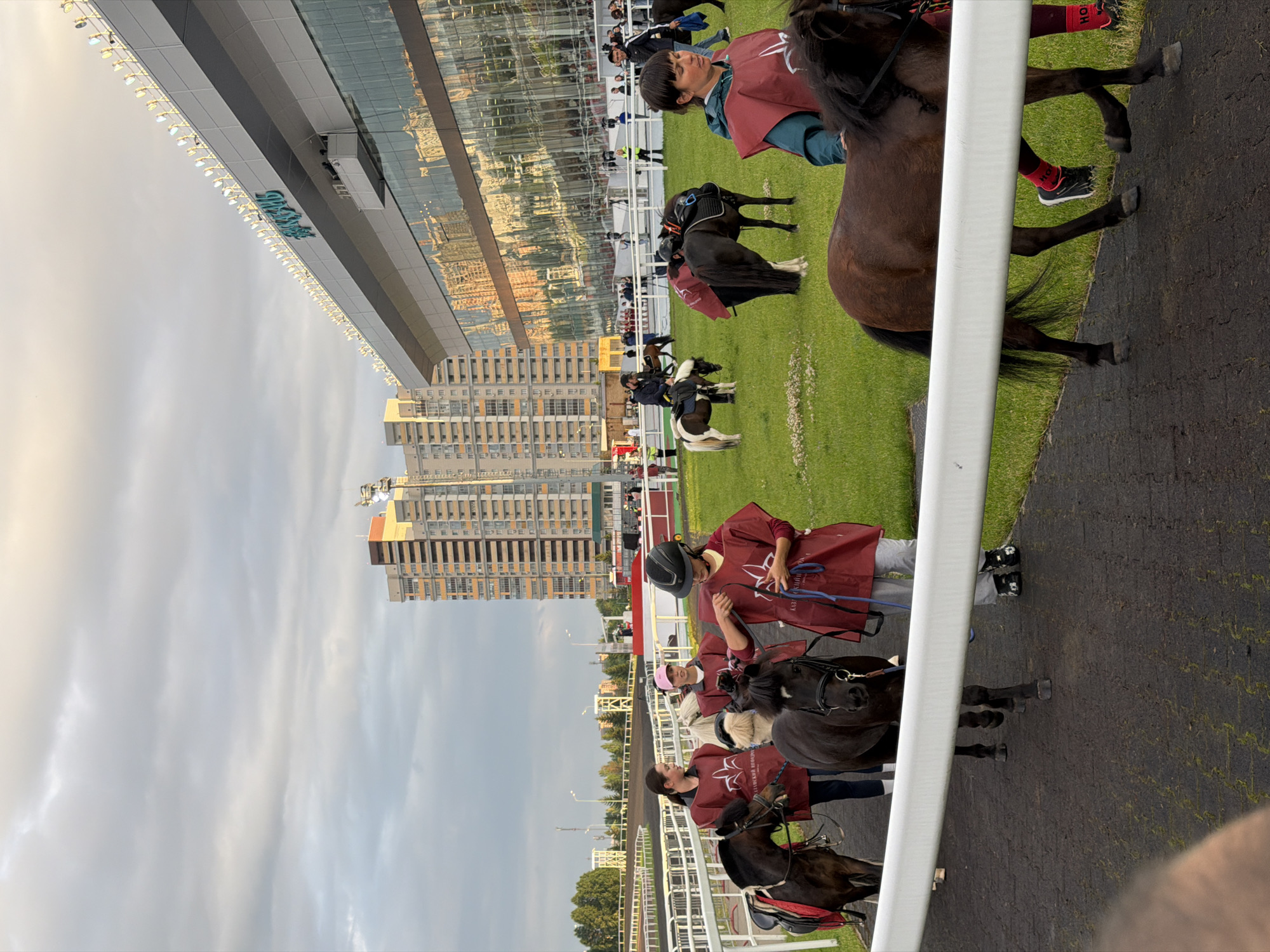In the city of Unknown, Unknown, a group of people are walking with miniature horses on a racetrack. The sky is cloudy and overcast, and the sun is setting, casting a warm glow on the scene. A large, modern stadium with a reflective glass facade dominates the background, with tall buildings visible behind it. The racetrack is lined with white fences, and the grass area is green and well-maintained. People are wearing dark red vests over their clothing, and some of the horses are wearing saddles and bridles. The overall impression is one of an organized event involving children and ponies.