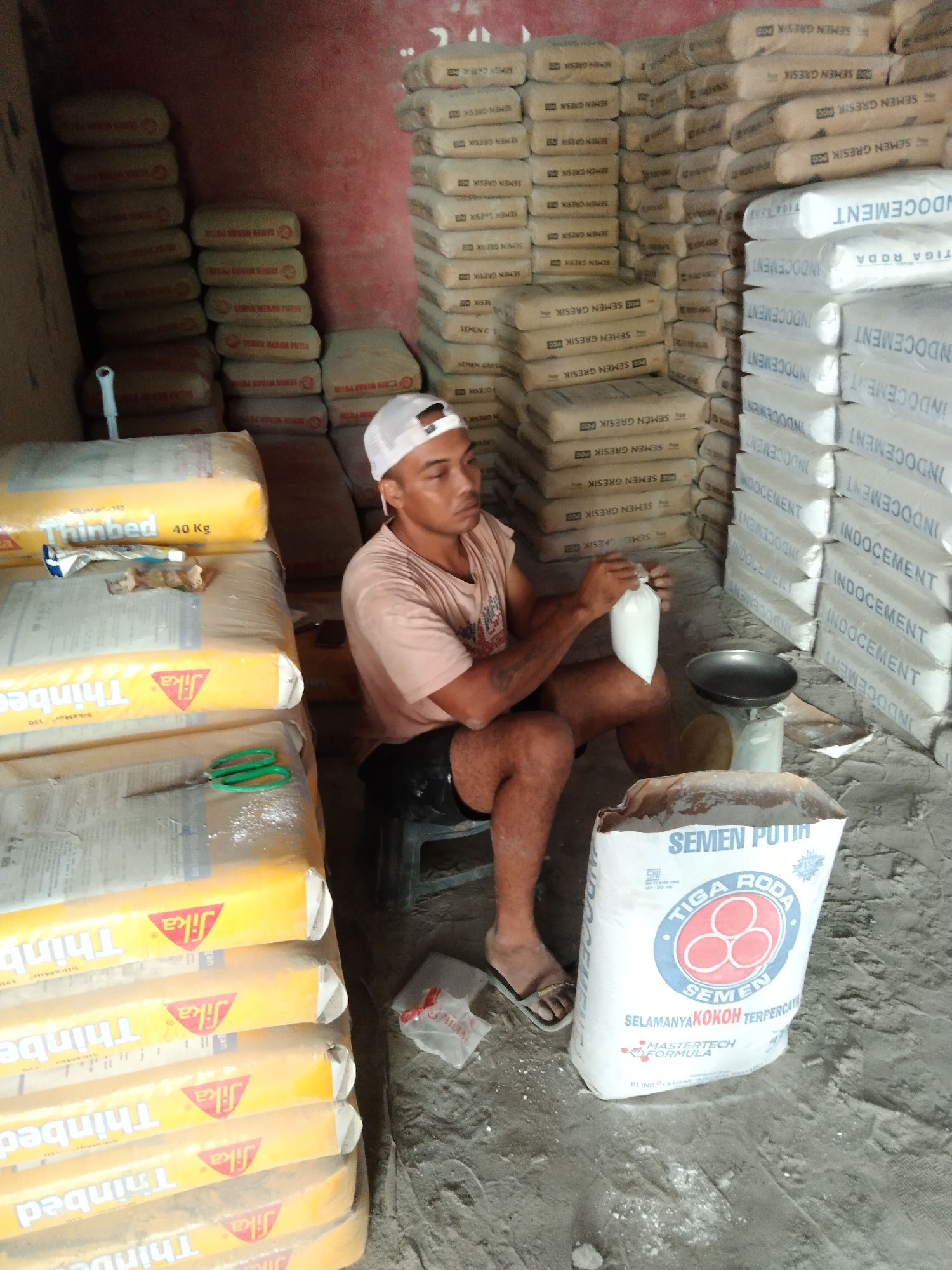 A man in a white cap and a light pink t-shirt sits on a stool in a storage room. He is holding a small white bag in his right hand, and his left hand is resting on a scale. In front of him, a large white bag with red and blue lettering is on the ground. The room is filled with stacks of bagged materials, some yellow and some light brown. The yellow bags have the brand "Thinbed" and "40 Kg" printed on them. The brown bags have various writings, including "SEMEN GRESIK" and "INDOCEMENT". A pair of green scissors is on a stack of yellow bags to the left. The floor is dusty and appears to be made of concrete. The overall scene suggests a place of business, likely a construction supply store or warehouse, in Parung, Indonesia.