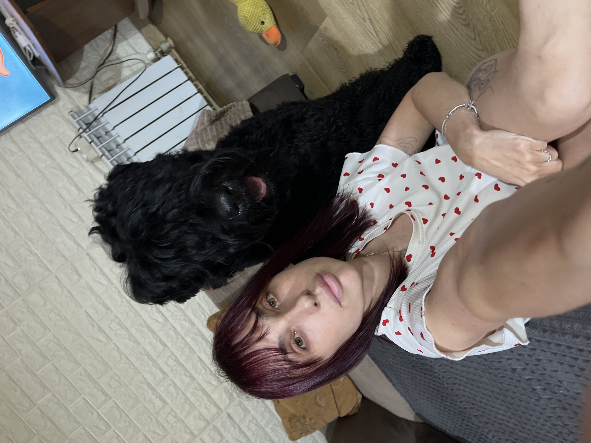 A medium shot shows a woman with dark red hair and bangs, wearing a white shirt with small red hearts, sitting on the floor next to a large black dog. The woman is looking towards the camera, with her head tilted slightly. The dog is behind her, with its mouth open as if panting or smiling. The background includes a white brick-patterned wall, a radiator, a TV screen, and a yellow duck toy. The floor is made of light brown wood. The scene appears to be indoors, possibly in a living room. The lighting is warm and natural. There is a couch to the left of the woman, partially visible.