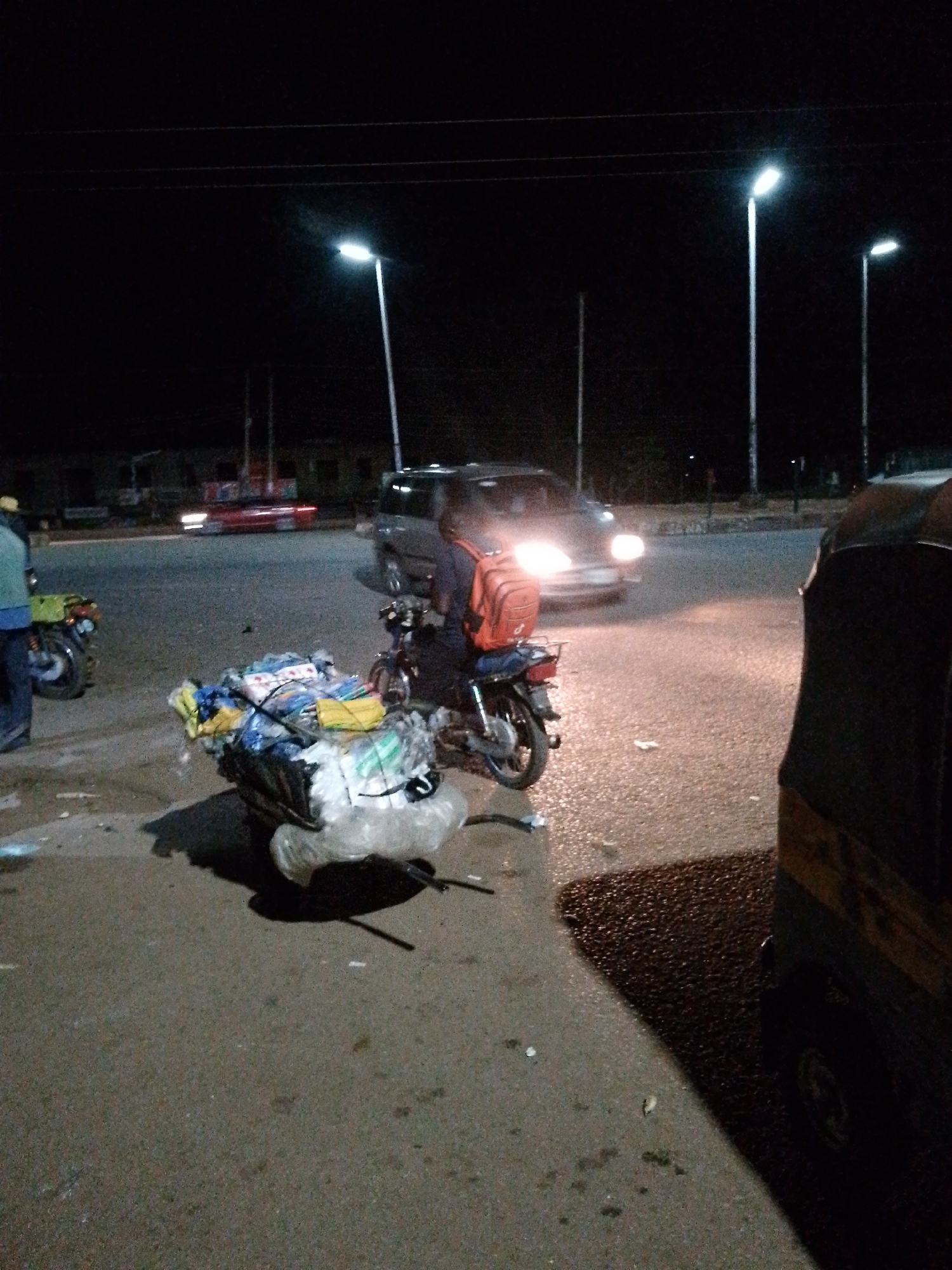 A nighttime street scene in an urban environment. A person is seated on a motorcycle, which is heavily laden with what appears to be personal belongings or goods wrapped in plastic and fabric. The motorcycle is parked on the right side of the frame, facing away from the viewer, with its rear wheel and cargo clearly visible. The cargo is a large, bulky bundle tied securely to the back of the motorcycle. Further down the street, a car with its headlights on is moving towards the viewer. In the foreground, to the right, a portion of a three-wheeled vehicle, possibly an auto-rickshaw, is visible. The scene is illuminated by streetlights and the headlights of vehicles, creating a dark, atmospheric setting. The road surface appears wet, suggesting recent rain. The overall impression is of a transport scenario at night, with the motorcycle and its rider preparing to move or having just stopped.