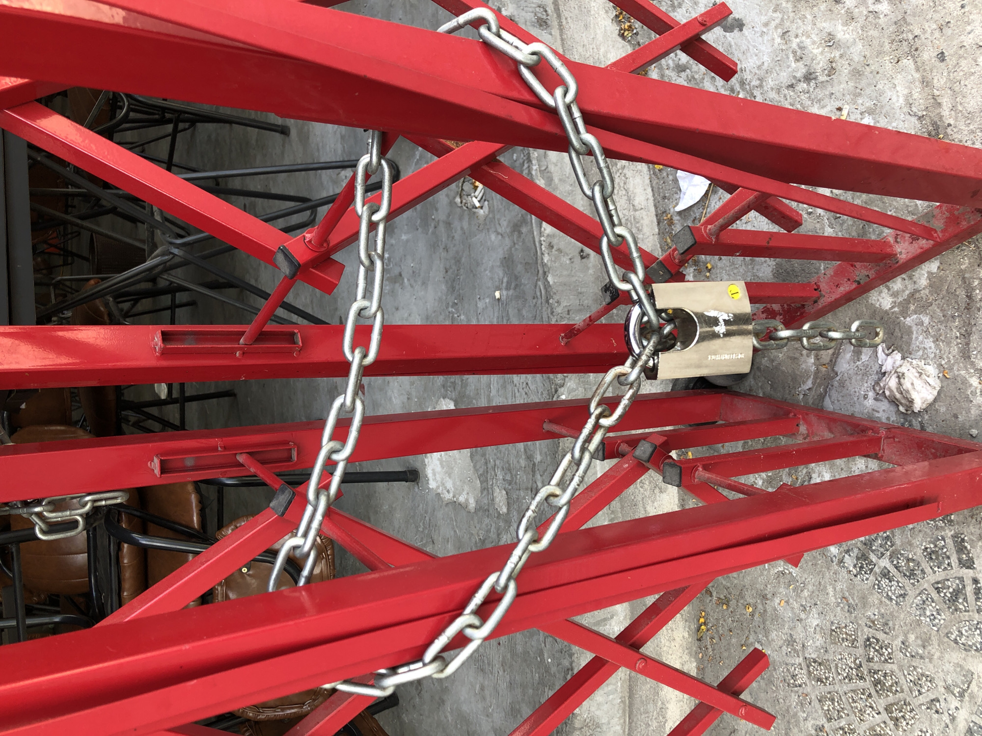 An outdoor scene in Da Nang, Vietnam, features a closed red metal gate, likely a storefront or entryway, secured with a silver chain and a silver padlock. The gate is made of interlocking red metal bars, forming an expandable barrier. The chain wraps around the gate structure and passes through the shackle of the padlock. The padlock itself is silver and appears to be a common brand, with some text visible on its face. Behind the gate, chairs and tables are visible, suggesting the space may be a restaurant or café. The ground is paved with concrete and small stones, and some debris, including fallen leaves and a white cloth, is present on the ground. The overall impression is one of closure or security.
