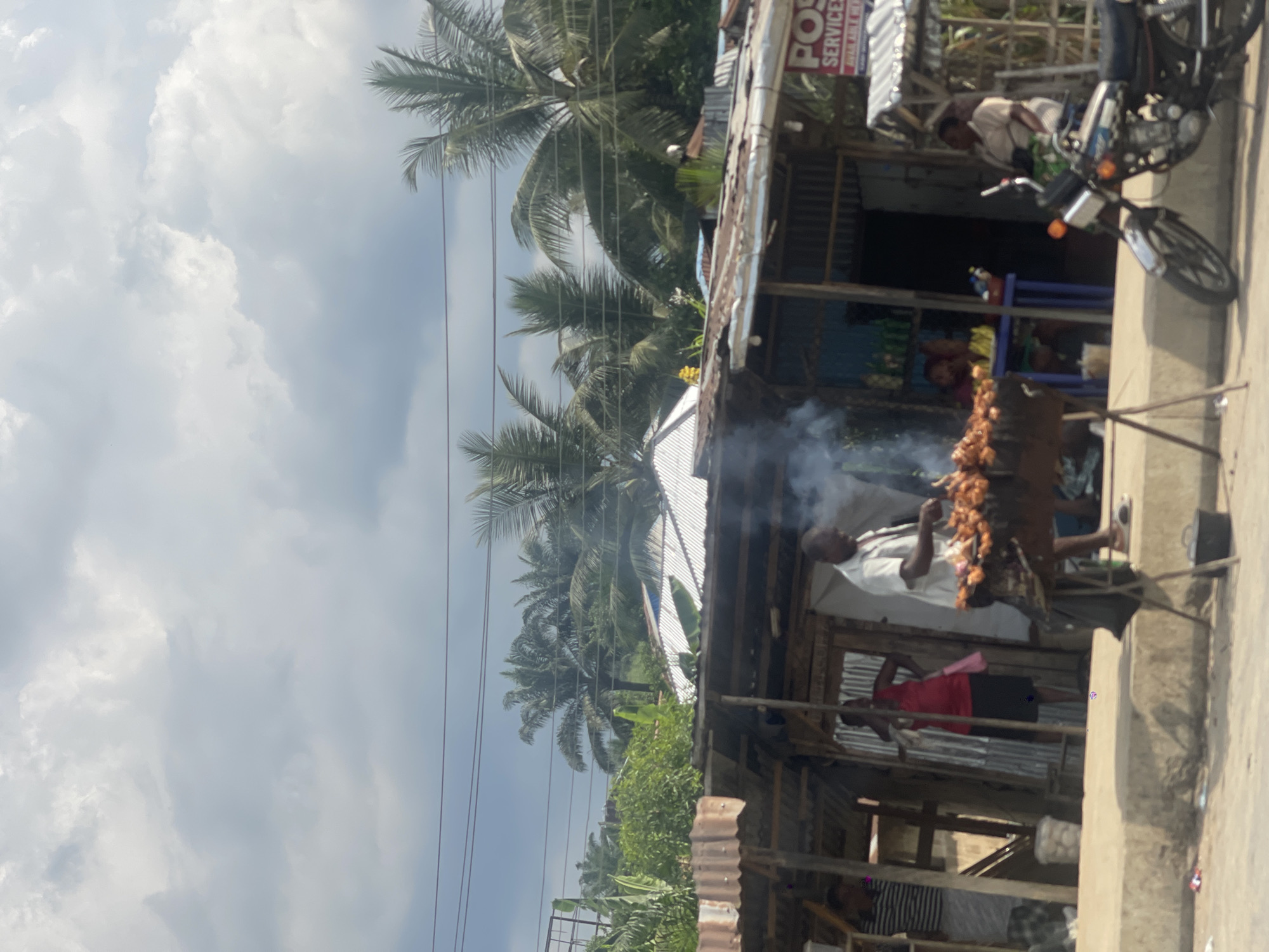 This outdoor image, rotated 90 degrees clockwise, captures a bustling roadside scene, likely in a tropical or subtropical region. In the foreground, a man in a light-colored shirt is actively grilling skewers over an open flame, producing a significant amount of white smoke. The skewers appear to be laden with pieces of meat or poultry. He stands before a rustic, open-fronted structure made of wood and corrugated metal, typical of a small shop or food stand.
Behind the griller, another person wearing a red top and dark skirt is partially visible inside the structure. Further inside, other individuals and various goods can be seen, suggesting a vendor's stall. A black motorcycle is parked prominently near the stand, adding to the street-level activity.
Above and behind the structure, lush green vegetation, dominated by numerous tall palm trees, extends across the midground, indicating a warm climate. Power lines stretch horizontally across the upper part of the scene, intersecting the sky and the palm fronds. The sky above is bright and heavily clouded with large, white cumulus clouds, though hints of blue sky are visible.
Notable details include a red and white sign affixed to the shop's roof, which partially reads "POS SERVICE" and "AVAILABLE HERE." The overall atmosphere suggests a vibrant, informal marketplace or a busy roadside food establishment during daylight hours.