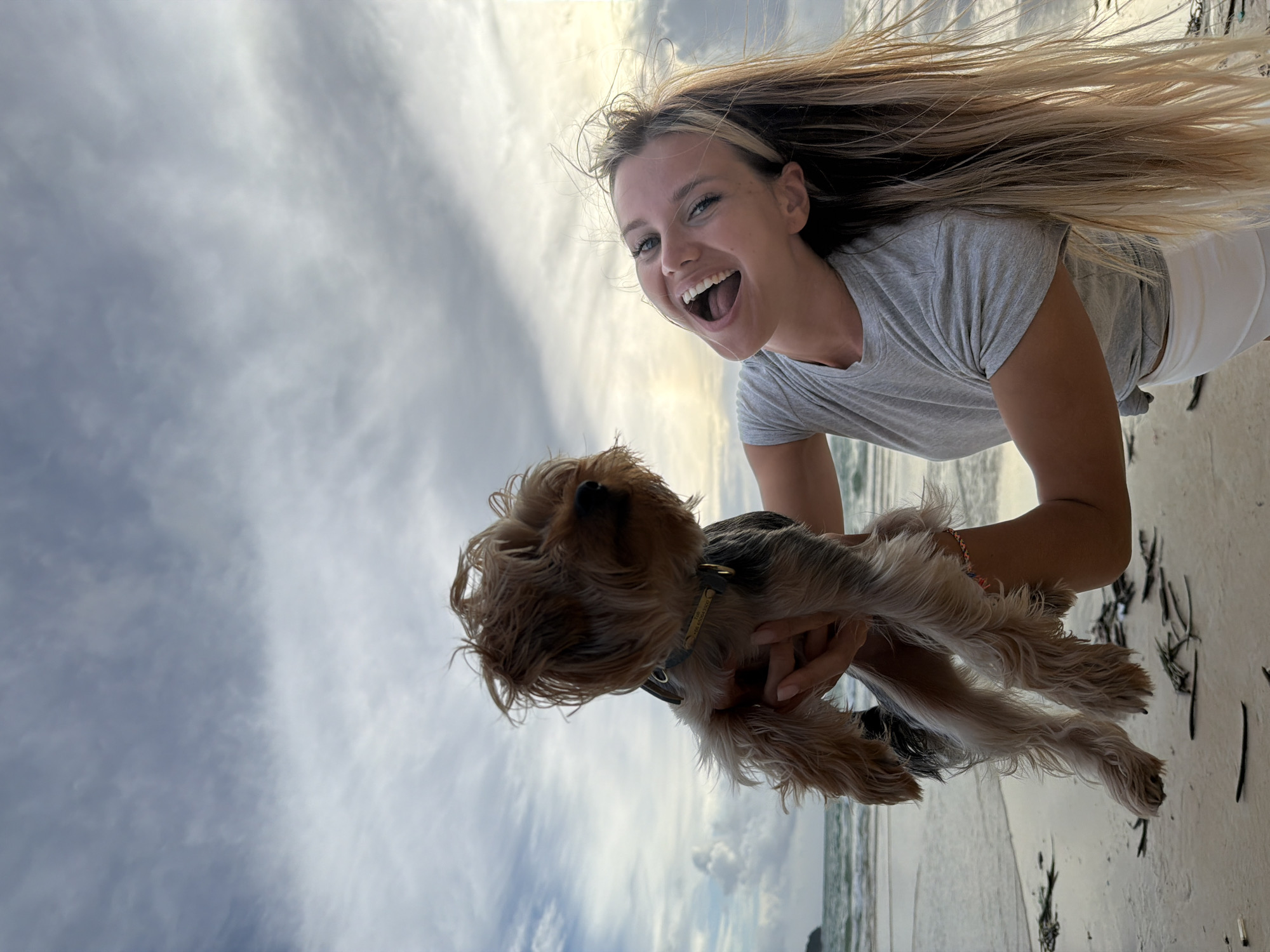 A young Caucasian woman with long blonde hair is holding a small, brown and grey Yorkshire Terrier on a beach in Sakhu, Thailand. The woman is wearing a grey t-shirt and white shorts, and she is laughing with her mouth wide open. The dog is being held up in the air by the woman. The beach appears to be sandy, with some debris on the ground. The ocean is visible in the background, with gentle waves and a cloudy sky. The lighting suggests it may be late afternoon or early evening.