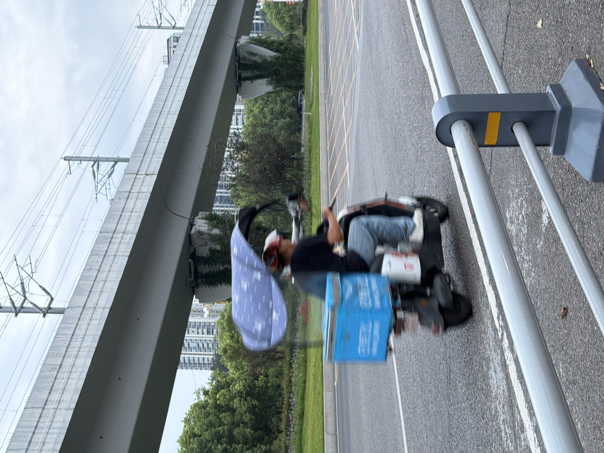 An individual on a scooter is captured in motion on a street in Jiangjin, China, beneath an elevated structure that appears to be a bridge or overpass. The scooter is equipped with a large, blue insulated box on its rear, suggesting it is used for deliveries. The scooter also has a canopy-like shield attached, patterned with a light blue fabric and white polka dots, likely to provide shade or protection from the elements. The person riding the scooter is wearing a dark shirt, blue jeans, and a helmet. The background includes lush green trees, distant urban buildings, and overhead power lines supported by poles. The street is paved, with lane markings visible, and a metal railing runs along the foreground. The sky is overcast.