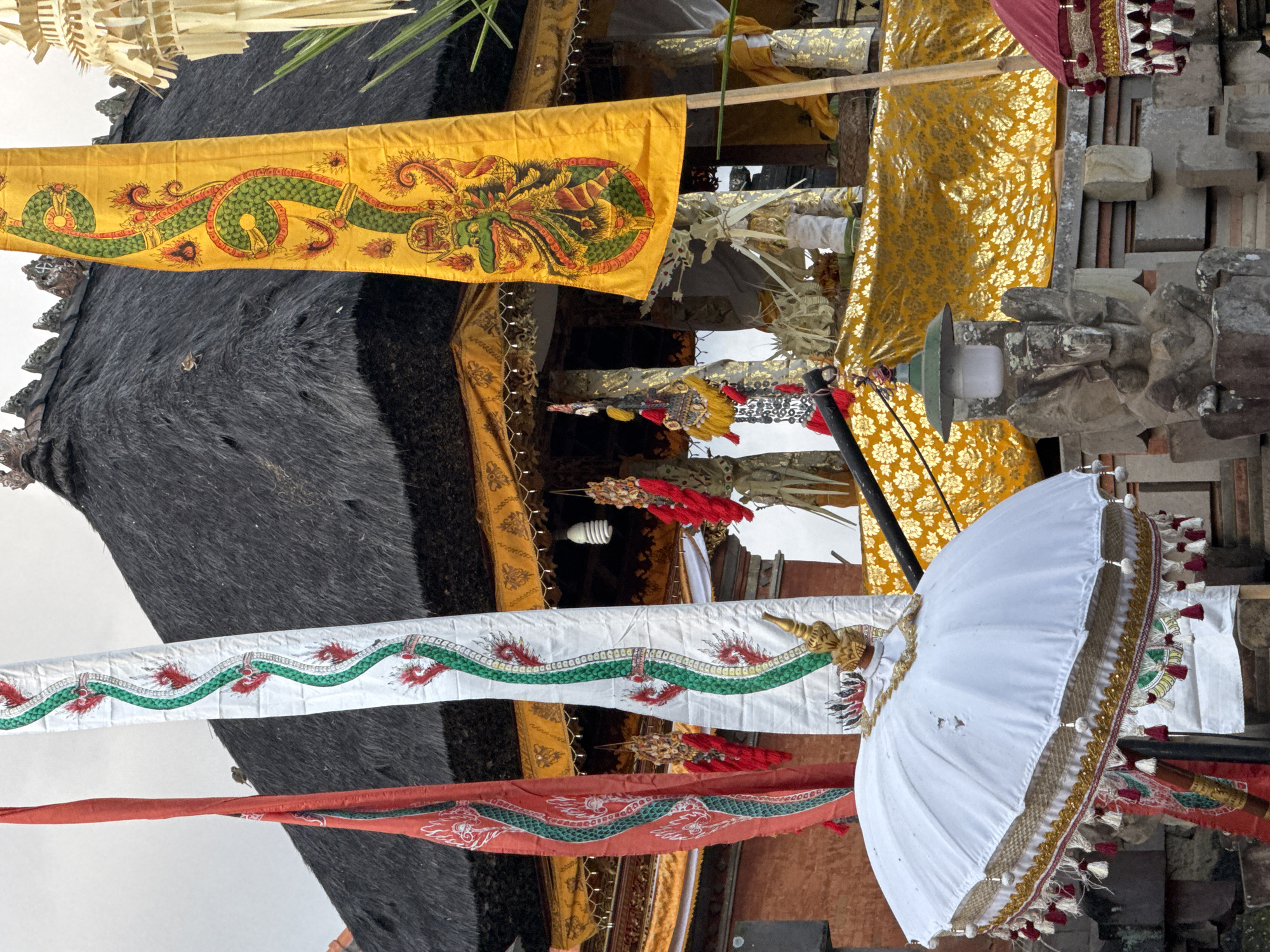 This is a medium shot, exterior photograph taken at the Pura Goa Gajah temple in Blahbatuh, Indonesia. The temple features a thatched roof and is decorated with several traditional Balinese banners and umbrellas. The banner on the right is yellow with a green and red dragon design, while the banner in the center is white with a green and red dragon design. A red banner is partially visible on the left. A large white umbrella with decorative tassels is positioned in the foreground, to the left of center. In the background, elaborate golden fabric adorns a shrine area. A stone statue with a lamp on top is situated in the lower right quadrant of the image, in front of the shrine. The sky is overcast. The overall scene depicts a cultural and religious setting.