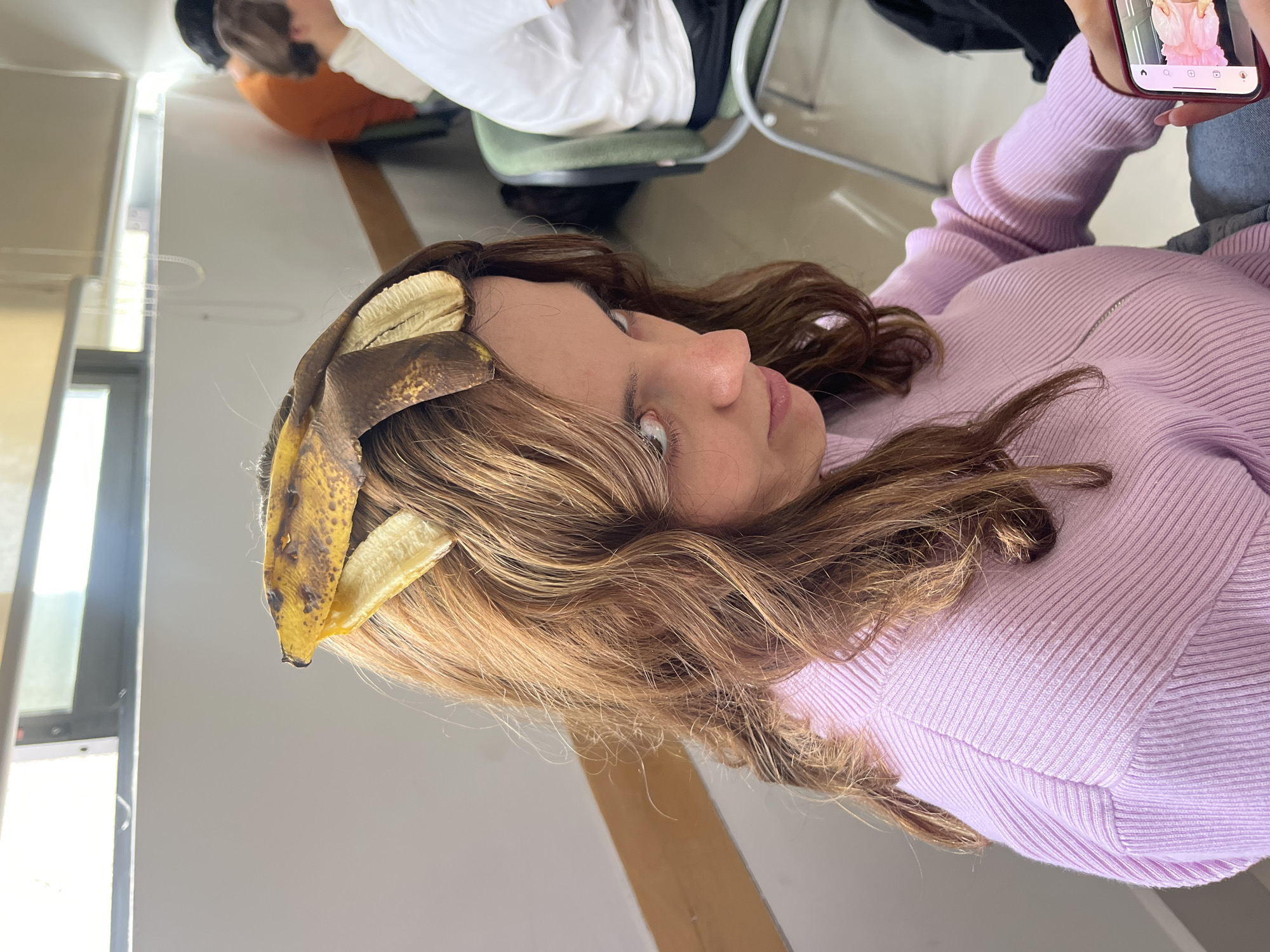 This indoor photograph, taken in San Andrés Cholula, Mexico, captures a young woman with a banana peel whimsically placed in her long, wavy brown hair. She is seen from a side profile, looking slightly upwards with a neutral expression. She wears a form-fitting, ribbed light purple long-sleeved top. Her right hand is holding a red-cased smartphone above her head, displaying what appears to be an Instagram feed with an image of a person in a pink ruffled top.
In the blurred background, other individuals are visible. One person wears a white top and a dark jacket, seated on a green office-style chair. Another person can be seen wearing an orange top. The setting suggests a casual environment, possibly a classroom or common area, with light-colored walls and flooring, and reflections indicating natural light from windows or bright overhead lighting. The activity seems to be a playful or candid moment, possibly captured during a break or informal gathering. Visible text on the phone screen includes Instagram interface elements like home, search, post, reels, and profile icons. The time of day appears to be daylight due to the bright ambient light.