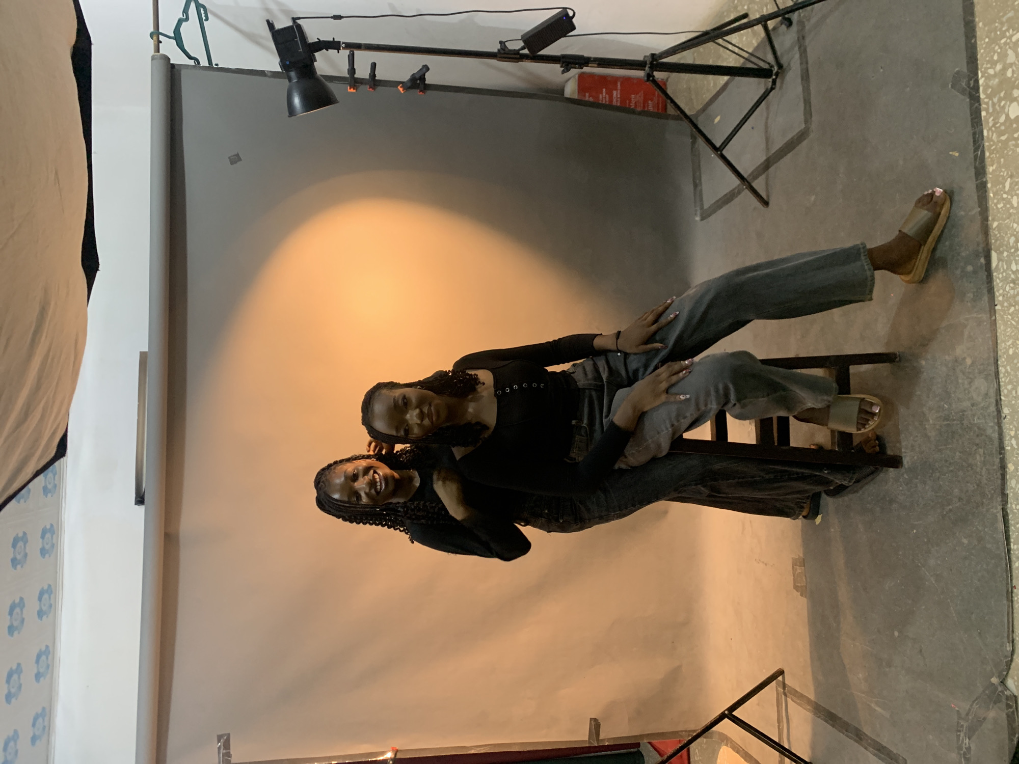 Two women are posing for a photograph in a studio setting. The studio is equipped with a backdrop, lighting equipment including a spotlight and light stand. The women are both wearing dark clothing. One woman is sitting on a chair. The location appears to be an indoor photography studio, not identifiable as being in Gura Top, Nigeria.