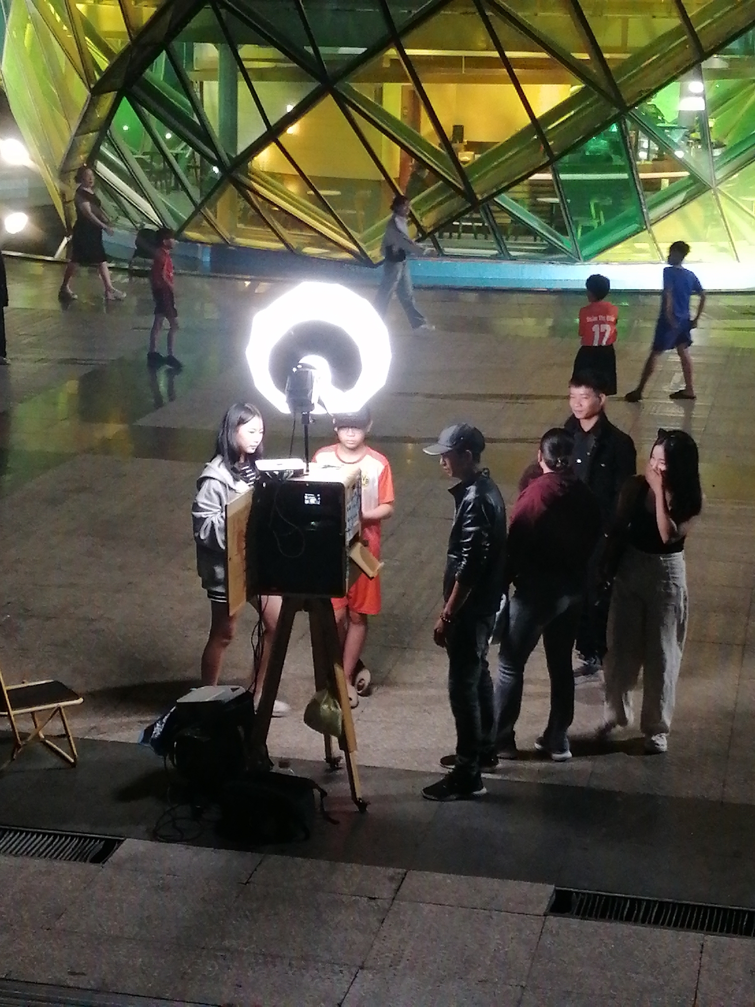 A group of people is assembled in front of a photo booth, illuminated by a ring light, likely in Da Lat, Vietnam, given the presence of the distinctive architectural structure in the background, resembling a large glass-covered geometric form. A young woman in a hoodie stands next to the photo booth with a boy in an orange shirt. There is a tripod supporting the camera and a screen to view the photos. A man in a leather jacket and a cap stands to the right. Behind him are two other people, one in a dark jacket. A woman in a black top and white pants stands near the last two people. In the background, other people are walking in front of the structure. The ground is paved. A black case and a folded chair are next to the photo booth setup.