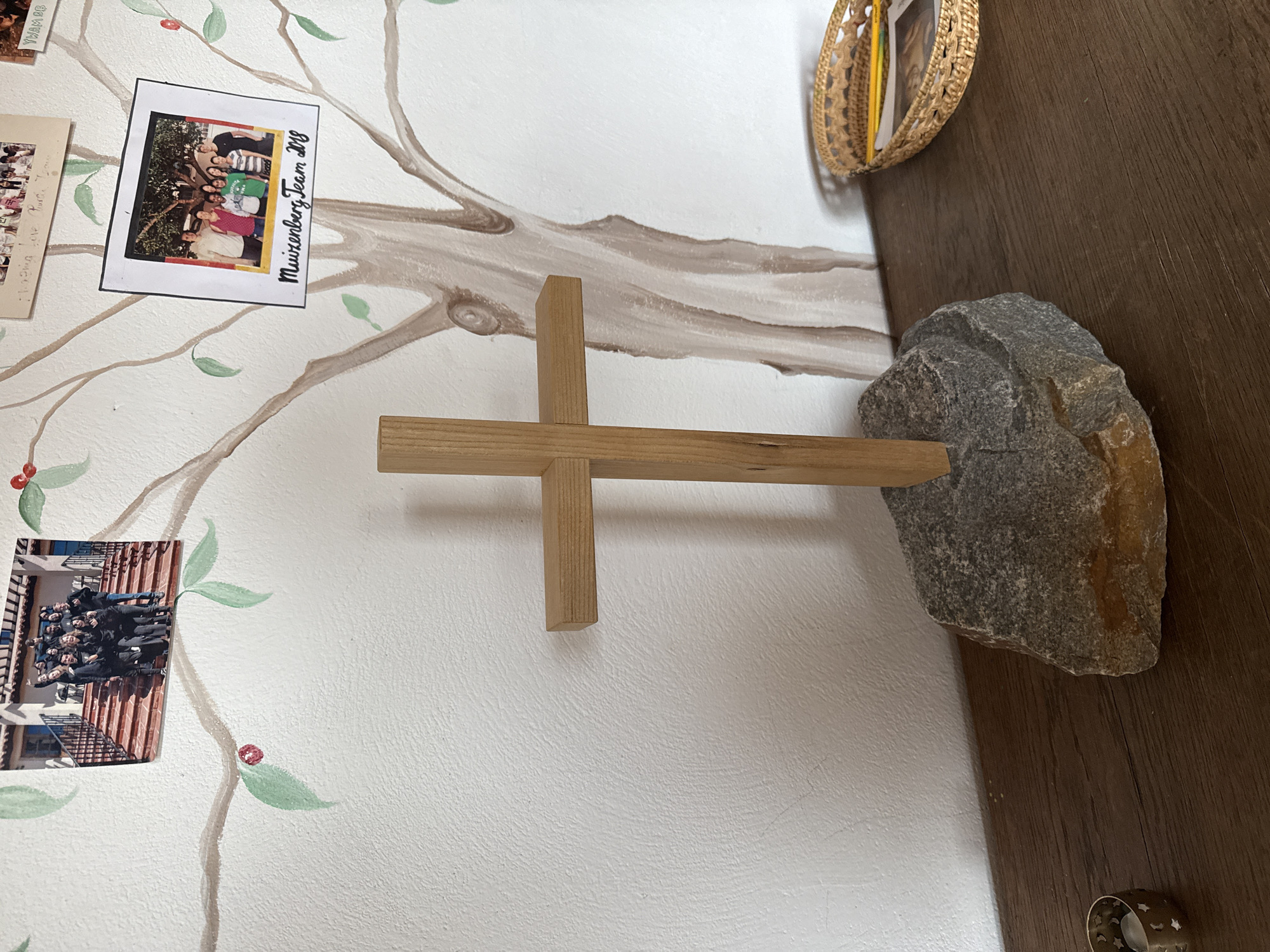 The image depicts a wooden cross standing on a large, rough stone. The cross is positioned in the foreground, centered on a dark wooden surface. Behind the cross, a white wall is visible, adorned with a mural of a stylized tree with brown branches and green leaves. Several framed photographs are attached to the mural. One photo, at the top left, shows a group of people on stairs in front of a building. Another photo, to the right, is labeled "Muiizenberg Team 2018" and shows a group of people standing under a tree. Further to the right, a woven basket contains pencils and what appears to be a photograph. A small, star-patterned candle holder with a lit candle is visible on the left side of the wooden surface. The scene appears to be an indoor setting, possibly a room in Bad Blankenburg, Germany, given the context provided.
