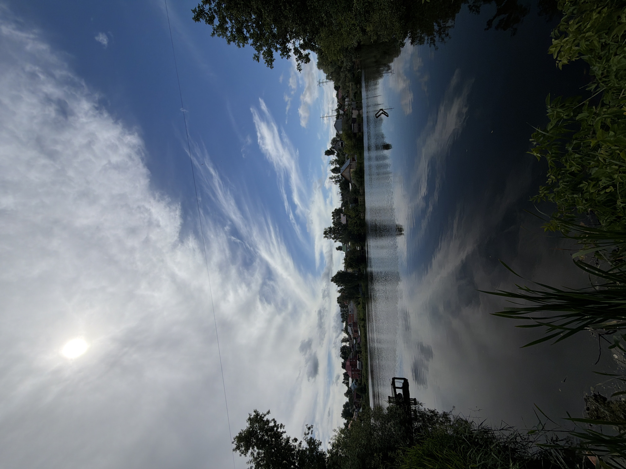 The image depicts a serene natural landscape with a body of water in the foreground, likely the Kama River in Chaikovsky, Russia, given the context of the provided cropped images. The water is calm, reflecting the sky and the surrounding environment. In the middle ground, there is a bank lined with trees and houses, suggesting a settlement or village. The sky is a vibrant blue with scattered white clouds, some of which are wispy and stretched across the frame. The sun is visible in the upper left corner, partially obscured by clouds, casting a bright glow. The foreground shows dense green vegetation, including tall grasses and bushes, growing along the edge of the water. There is a small wooden dock on the left side of the image, extending into the water. The overall impression is one of a peaceful, rural or semi-rural setting on a clear day.