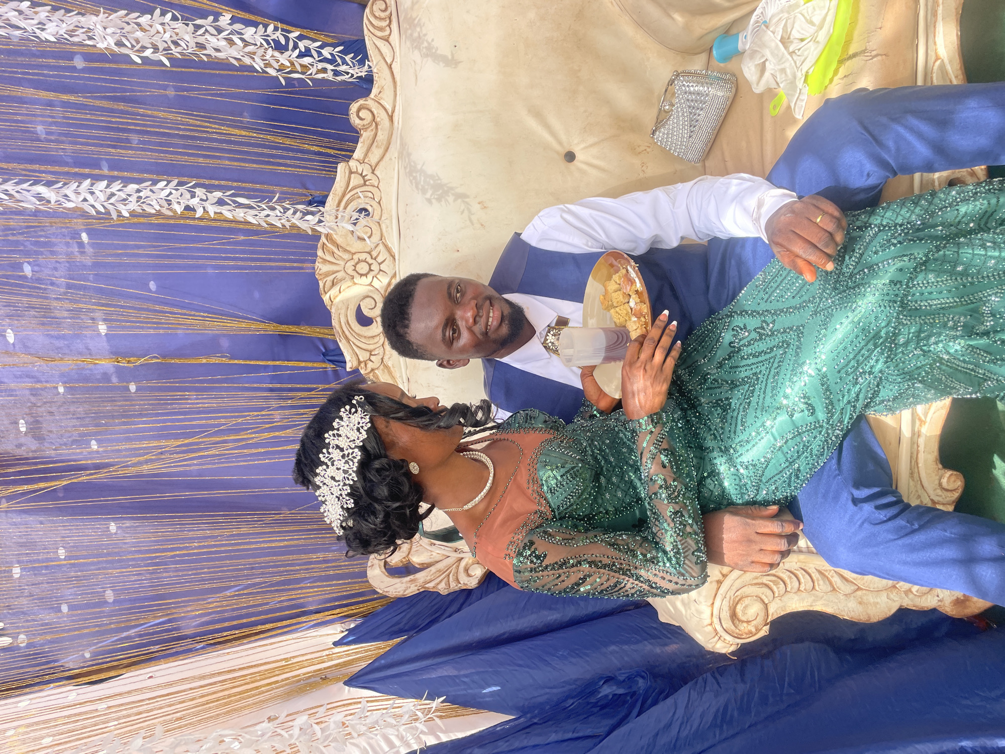 A couple is seated on an ornate, beige couch, likely during a wedding celebration in Ogbe, Nigeria. The man, dark-skinned and wearing a blue suit jacket over a white shirt and blue pants, is holding a plate of food and a clear cup. He is looking towards the woman with a smile. The woman, also dark-skinned, wears a green, sequined gown and a decorative headpiece. She is facing the man. The background consists of blue fabric adorned with gold tinsel and white floral garlands. A silver clutch purse is on the couch near the man.