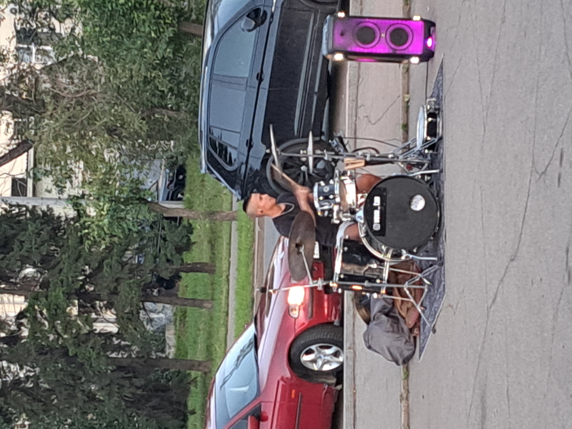 The image shows a street musician playing a drum set on a city street. The musician, a young man with a mohawk hairstyle, is seated behind his drum kit, holding drumsticks and actively playing. The drum set includes various drums and cymbals, set up on a patterned mat on the asphalt. To the right of the drum set, a portable speaker with purple lighting is visible. In the background, there are parked cars, including a red car on the left and a black SUV on the right. Trees and a building can be seen behind the cars. The scene appears to be outdoors during daylight. The musician is positioned in front of a red car, and a black SUV is parked behind him. The overall setting suggests a public street in an urban environment, likely a pedestrian area or a less busy section of road, where street performances might take place. Based on the provided context, the location is identified as the city of Irkutsk, Russia.