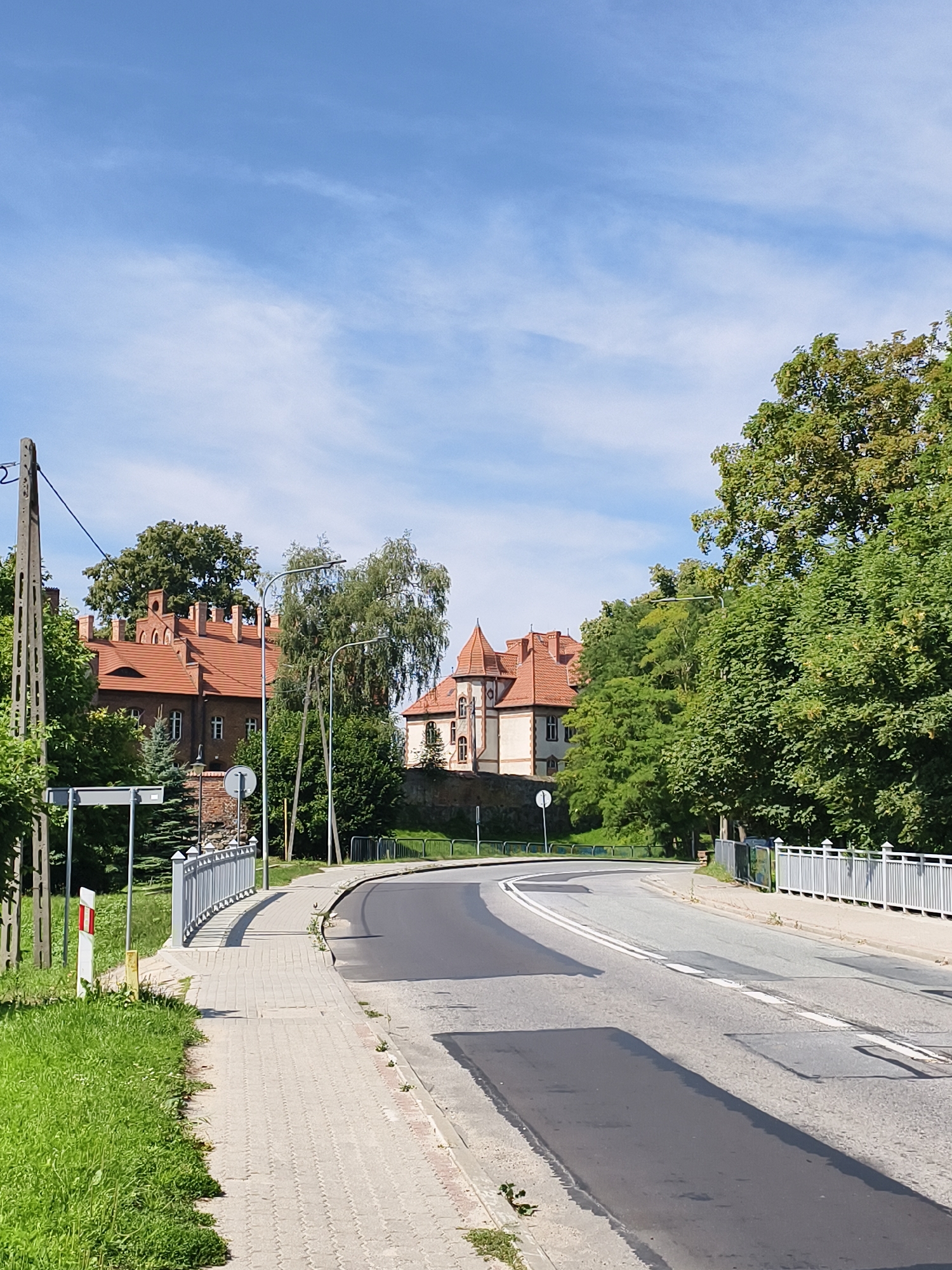 The image captures a street scene in Sztum, Poland, on a clear, sunny day with a blue sky dotted with wispy clouds. The foreground shows a paved sidewalk running alongside a grassy verge, leading towards a curved road. The road itself is asphalt, marked with white lines, and appears to have undergone recent repairs with dark patches. A white metal railing delineates the sidewalk from the road on the right.
Further down the road, on the left side, a traditional brick building with a red tiled roof and several chimneys is visible, partially obscured by lush green trees. To the right of this building, a second, lighter-colored building with a distinctive turret and red tiled roof is also present, set behind a low stone wall. Streetlights are positioned along the sidewalk. The overall impression is a quiet, European town street lined with historic architecture and abundant greenery.