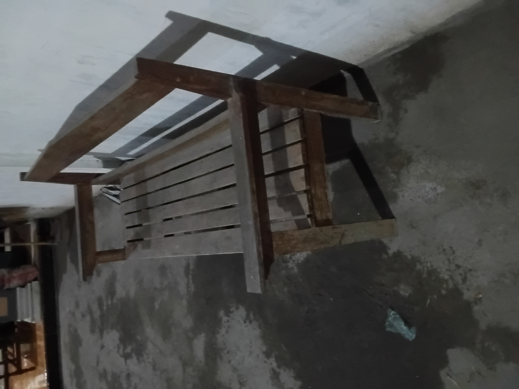 A wooden bench is placed against a light-colored wall. The bench is constructed of horizontal wooden slats for the seat and a solid wooden backrest. The floor is concrete and appears damp in patches, with some water stains and a crumpled piece of light-colored plastic visible to the left of the bench. Shadows from the bench and possibly other unseen objects are cast on the wall and floor. The lighting suggests an indoor or sheltered outdoor space, possibly a patio or covered walkway. There are no people or animals visible in the image.