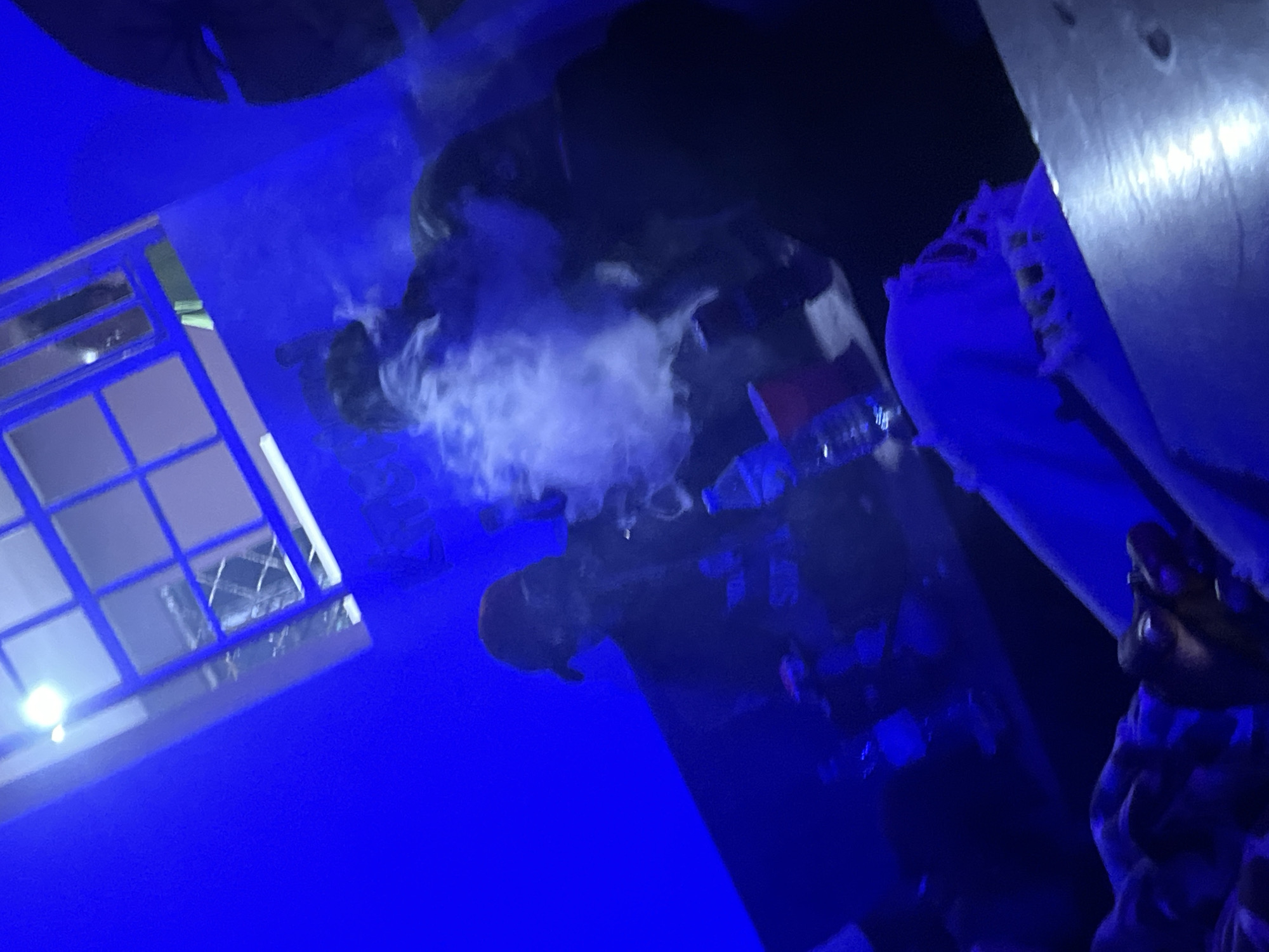 A dimly lit scene, bathed in blue light, depicts several individuals in what appears to be a social gathering. In the center, a person wearing a red cap and dark clothing is visible, exhaling a cloud of smoke, likely from a vape or hookah, which billows around them. Another person is partially visible to the right, also appearing to be engaged in smoking. In the foreground, the edge of a table is visible, with what looks like a person's hands resting on it, covered in patterned fabric. Several bottles and cups are scattered on surfaces, hinting at a relaxed, informal atmosphere. The environment includes a textured ceiling with what appears to be a window or light fixture, casting bright light. The overall ambiance suggests a nighttime event in a club or private residence. The location is specified as Unknown, Nigeria.
