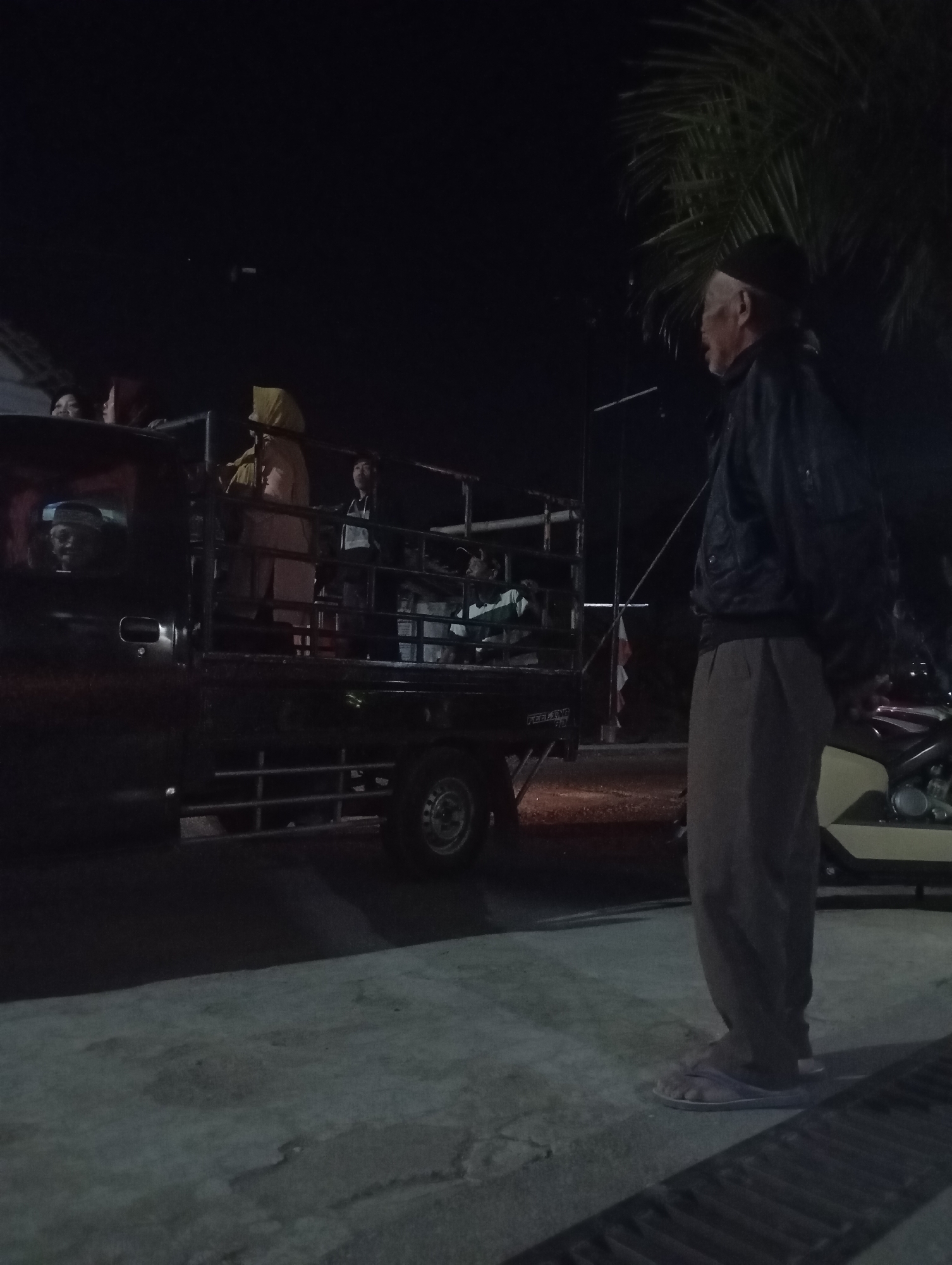 In Banjarsari, Indonesia, a man wearing a dark jacket, loose-fitting pants, and a skullcap stands on a paved surface at night, facing away from the camera towards a truck. The truck is carrying several passengers, some of whom are visible in the open bed of the vehicle. One passenger wears a yellow headscarf. The scene is illuminated by artificial light, casting shadows. A palm tree with dark fronds is visible against the night sky behind the man. A motorcycle is partially visible to the right of the frame. The overall impression is a snapshot of a community gathering or transportation event in an outdoor, dimly lit setting.
