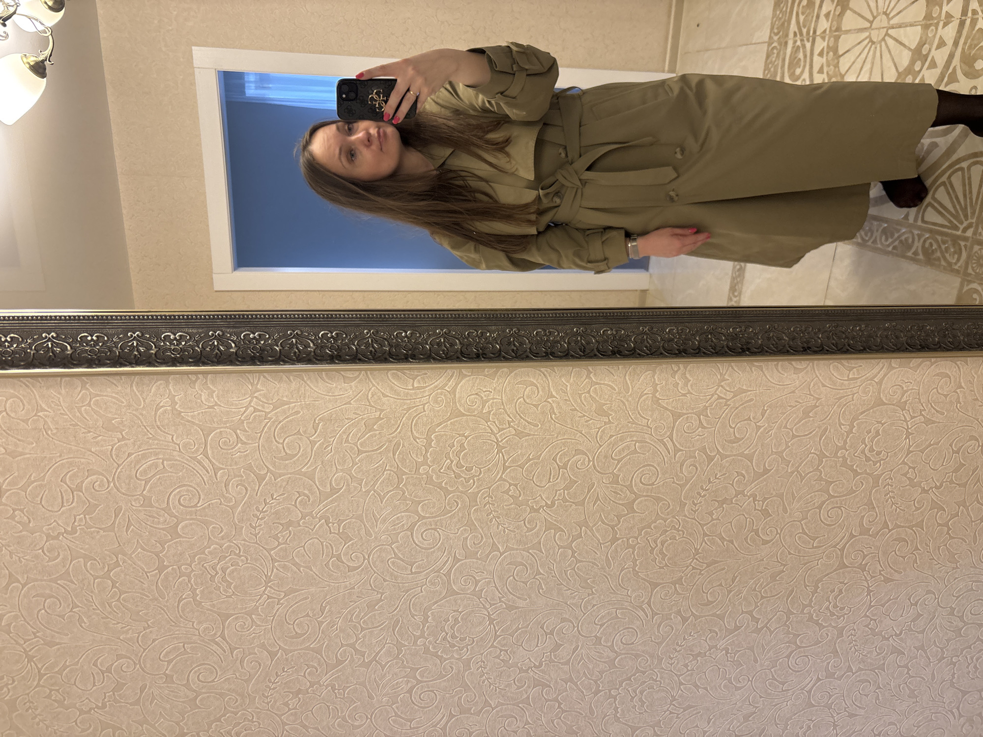 A woman is captured taking a full-body mirror selfie in an indoor setting, identified as Chaikovsky, Russia. She stands upright, holding a black smartphone with a "GUESS" branded case in her left hand, which is adorned with pink nail polish and a silver bracelet. Her long, straight brown hair falls over her shoulders. She is wearing a long, khaki or olive green trench coat with a tied belt, and dark tights or stockings are visible below the coat's hem.
The large mirror, featuring an ornate, dark-colored frame, reflects the woman and the room behind her. In the reflection, a white door frame is visible, beyond which a blue-toned wall or room is seen. The floor in the reflection is covered with decorative, light-colored tiles featuring intricate patterns.
To the left of the mirror, a light fixture with two frosted glass shades and brass accents is mounted on the wall. The wall surrounding the mirror is adorned with light beige or cream-colored wallpaper, embossed with a floral and scrollwork pattern. The woman's expression is neutral as she looks towards her phone.