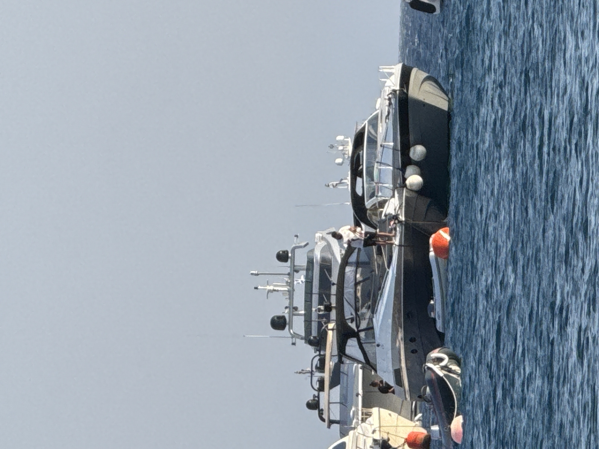 A view of a harbor in Agios Ioannis Diakoftis, Greece, with several yachts moored in the blue water under a clear sky. Two men are visible on the deck of a large, dark yacht in the foreground. One man, wearing a white shirt and dark shorts, stands near the stern. Another person, wearing dark clothing, is visible further forward on the same yacht. Several smaller dinghies and buoys are floating in the water around the yachts. In the background, a larger yacht with a white hull and dark accents is docked behind the foreground yacht. The overall scene depicts a calm day in a marina.