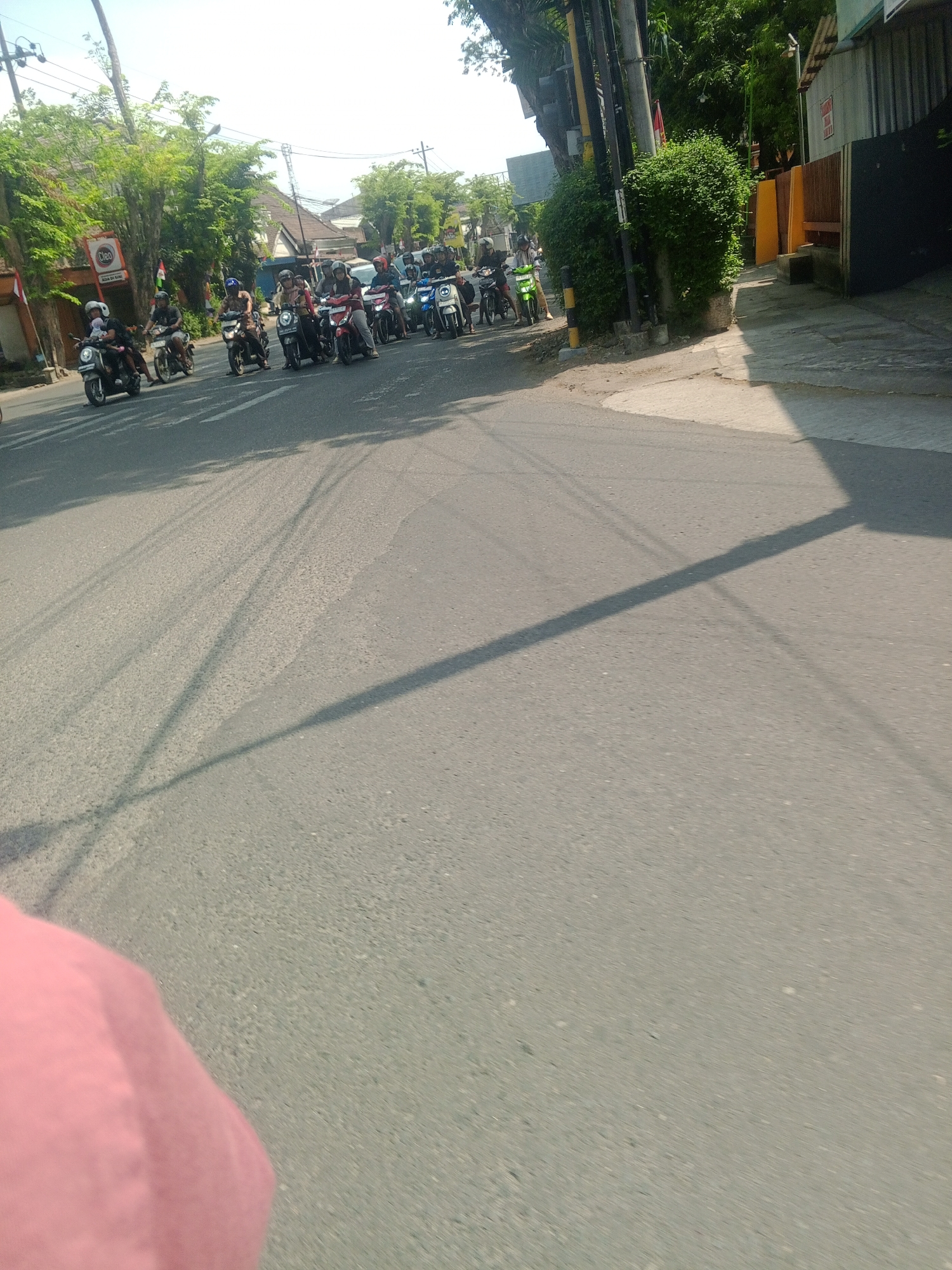 The image shows a group of people riding motorcycles on a road in Banyudono, Indonesia. The riders are mostly young adults and appear to be participating in a group ride or event. The environment is a typical Indonesian street scene with buildings, trees, and power lines visible. The road surface is asphalt. The lighting suggests it is daytime with bright sunlight, creating distinct shadows. One person in the foreground is wearing a pink garment, possibly a shirt or jacket. The motorcycles are of various models and colors, and some riders are wearing helmets. The overall atmosphere appears casual and social.