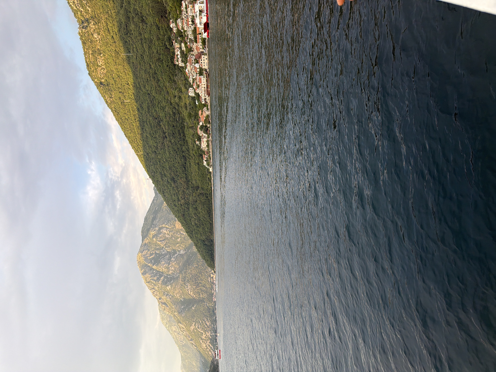 The image depicts a serene view of the Bay of Kotor in Lepetane, Montenegro, at golden hour. The foreground is dominated by the dark blue, slightly rippling water of the bay. In the mid-ground, a small settlement of white and red-roofed houses is nestled on the green, tree-covered slopes of a steep mountain that rises dramatically from the water's edge. The mountain's peak is partially illuminated by the setting sun, casting a warm glow on its rocky surface. A red and white ferry is visible on the water, approaching the shore, and another smaller white boat is seen further in the distance. The sky above is a mix of soft gray clouds and patches of blue, with the warm light of the sunset filtering through. The overall scene is tranquil and picturesque, characteristic of a Mediterranean coastal landscape.