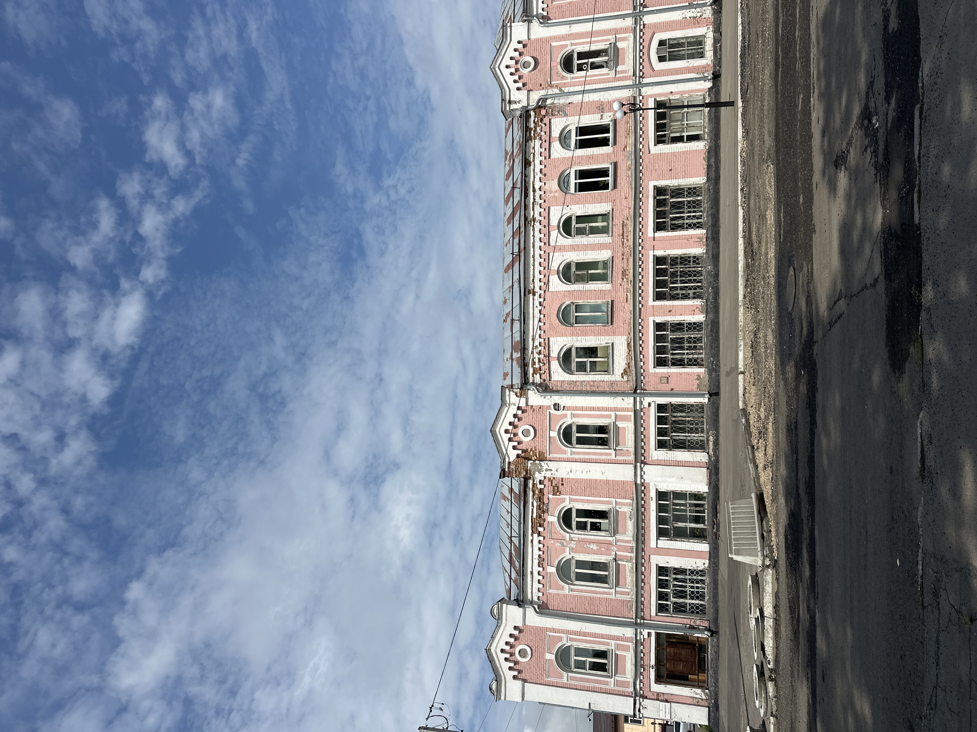 The image depicts a two-story, pink brick building with white decorative trim under a bright blue sky with scattered clouds. The building appears aged, with some sections of the facade showing signs of wear and damage, particularly along the roofline. It features numerous arched windows on the upper floor and rectangular windows with decorative metal grates on the lower floor. A dark wooden door is visible on the far left of the building's ground level. In front of the building, there is a paved road with some debris and construction materials, including concrete barriers and manhole covers. A single streetlamp stands near the center of the building's frontage. The scene suggests an urban or semi-urban environment, possibly in a historical district, located in Nogornyi, Russia. There are no people or discernible activity in the image.