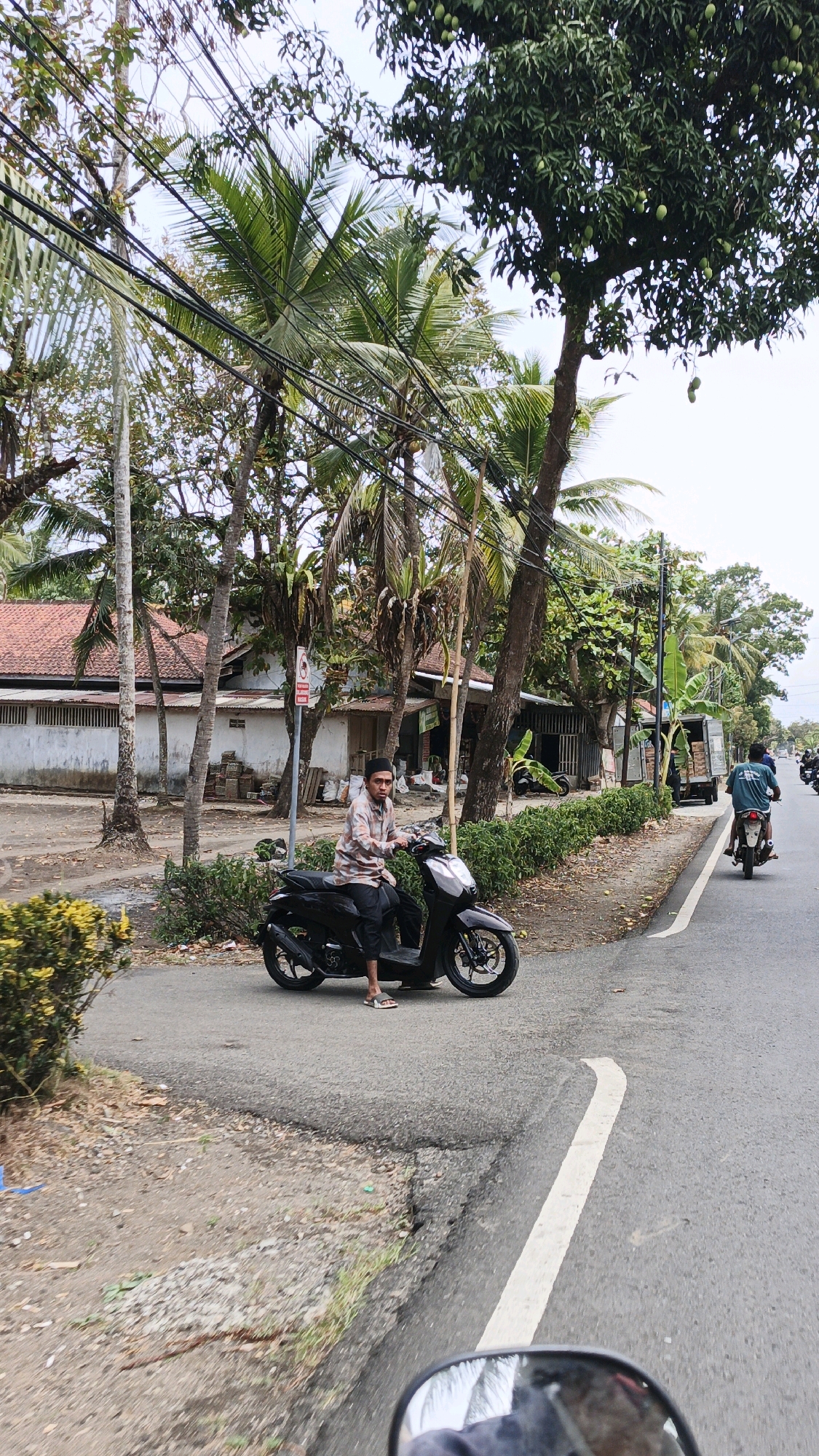 In Cibenda, Indonesia, a man wearing a checkered shirt and a peci cap sits on a black scooter on the side of a paved road. He is positioned on the left side of the frame, near a patch of yellow flowers. The road has a white lane marker. Behind him, a white building with a tiled roof is visible, with various stacks of materials in front. Palm trees and other foliage line the street, with power lines running overhead. Further down the road, another person on a scooter is visible, along with a truck. The sky is overcast.