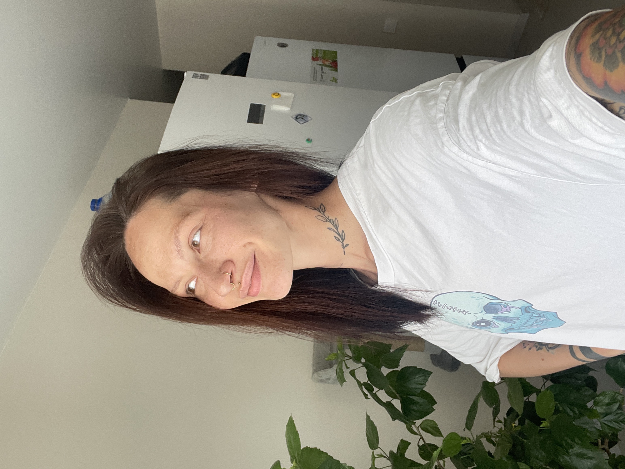 A person with brown hair, a nose ring, and tattoos on their neck and arms is wearing a white T-shirt with a skull graphic. They are positioned indoors near a green leafy plant. In the background, a white refrigerator is visible. There is no indication in the image or prompt that specifies the location is in the city of Izhevsk, Russia.
