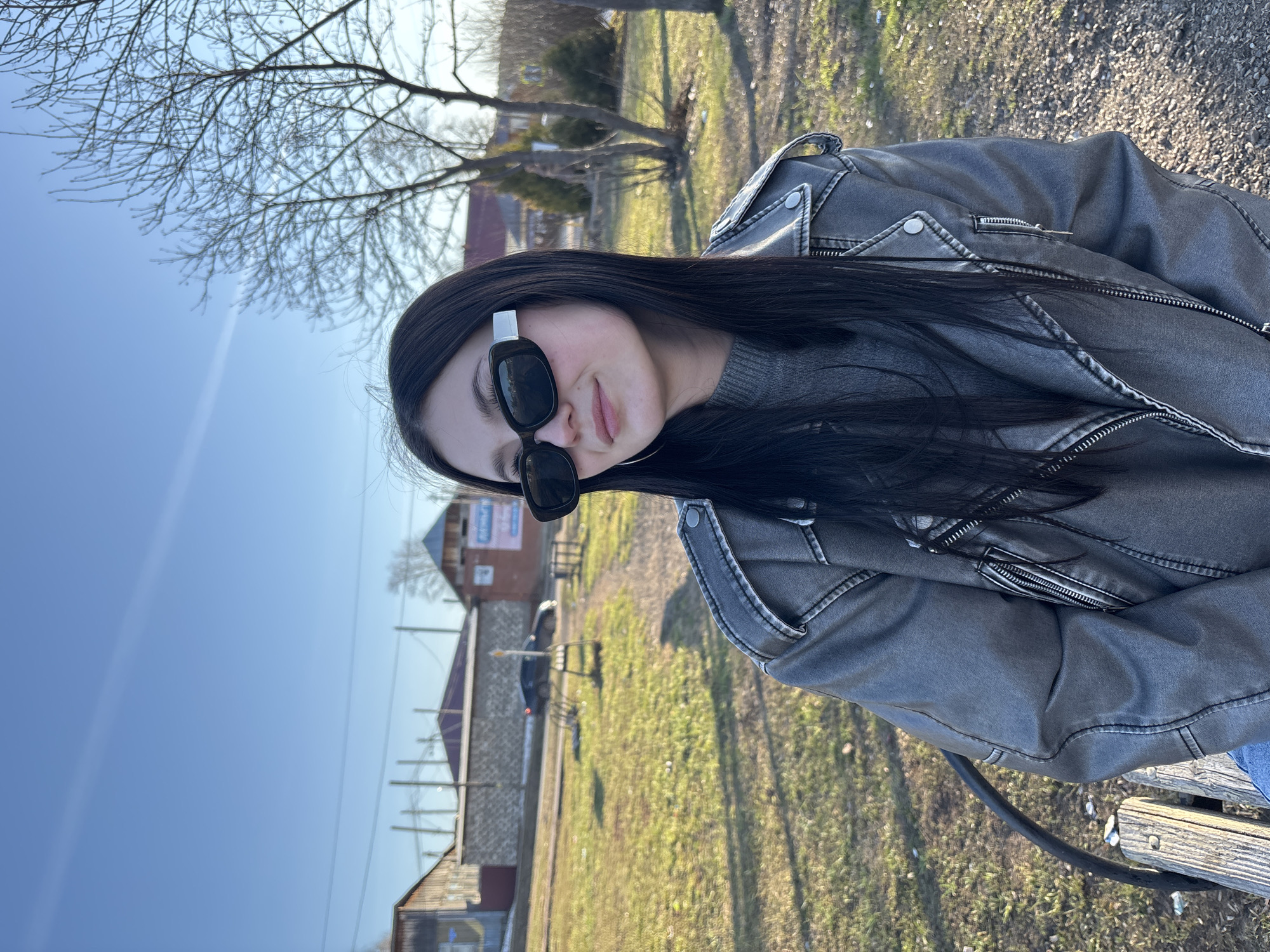 A young woman with long, straight dark hair is captured in a daylight outdoor shot, seated and looking towards the camera with a subtle, relaxed smile. She wears black sunglasses featuring a distinctive white or silver detail on the temples. Her attire consists of a grey ribbed sweater underneath a dark grey jacket, which appears to be made of textured material, possibly faux leather or distressed denim. The jacket is detailed with prominent stitching, silver snap buttons on the lapel, and several zippers, including one on a chest pocket and the main closure.
She is seated, likely on a wooden bench, with a portion of it and a black metal bar visible in the lower right of the frame. The setting is a bright, clear day, indicated by the vibrant blue sky that occupies a significant part of the background. Bare deciduous trees with intricate branch patterns stand against the sky, suggesting either late winter or early spring. The ground is covered with green grass, interspersed with areas of dirt or dry foliage. In the mid-ground and distant background, several buildings, resembling houses or rural structures, are scattered across the landscape. The overall impression is a calm, sunny day in a rural or semi-rural area. There is no clearly legible text visible in the image.