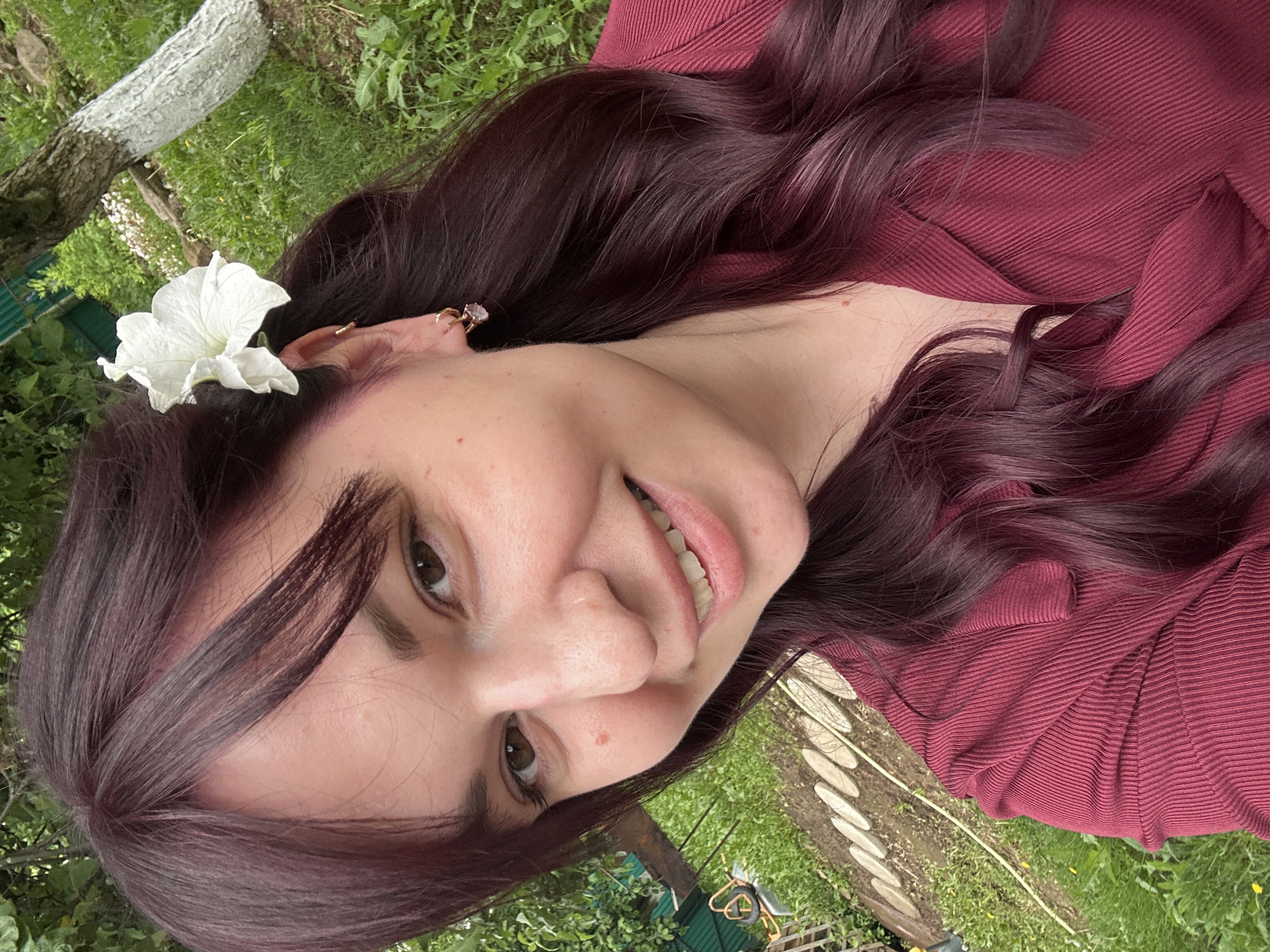 An adult female subject is depicted in a close-up, outdoor selfie taken in a garden setting within the city of Фоки, Russia. She has long, wavy, burgundy-colored hair with bangs across her forehead, and a white petunia flower is secured behind her right ear. Her right ear features multiple gold-toned earrings, including small hoops and a dangling earring with a pink gem. She has brown eyes, a mole on her right cheek, and is smiling with teeth visible, looking directly at the camera. Her attire consists of a deep red, ribbed, long-sleeved collared shirt. The background displays varied green foliage, a segment of a white-painted tree trunk on the right, and a garden path constructed from light-colored, oval stepping stones on the lower left. A dark green corrugated metal fence and a partially visible garden tool are also present in the background.