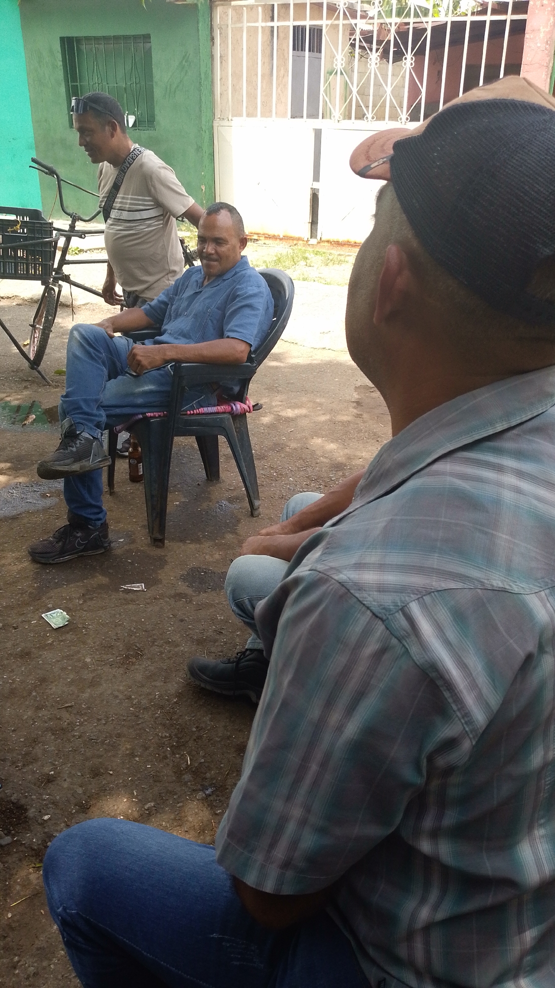 The image shows three men outdoors in Tinaquillo, Venezuela. One man sits in a black plastic chair wearing a blue shirt and jeans with black sneakers, his leg crossed over the other. A brown glass bottle sits on the ground near the chair. A second man, wearing a tan t-shirt, black glasses, and a black bag, stands behind the seated man, near a black bicycle with a basket. The third man, with a baseball cap, faces away from the camera wearing a blue and grey plaid shirt. The setting appears to be on dirt ground. There is a green house behind the first and second men and a white gate behind them.
