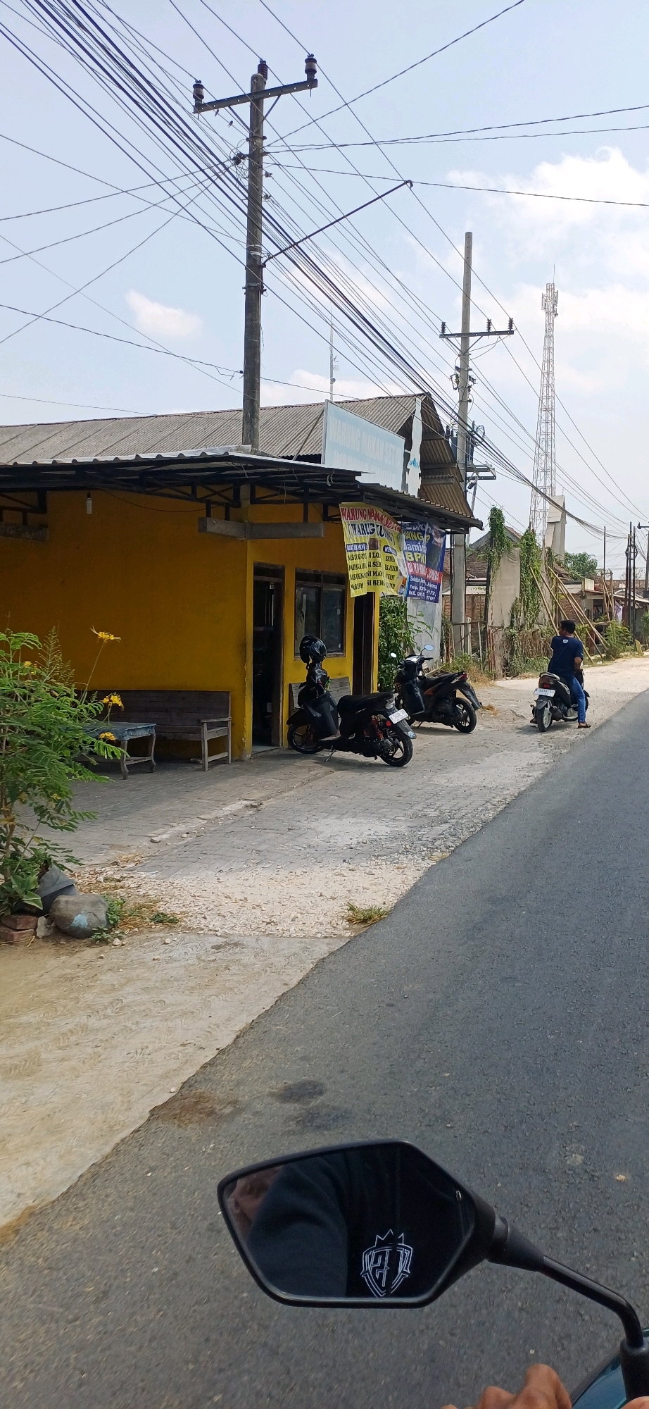 The image captures a street view in Jakenan, Indonesia, on a clear day with a light blue sky and scattered clouds. The scene features a small, yellow building with a corrugated metal roof and a covered porch. Various signs, some with Indonesian text like "WARUNG MAKAN SETU" and "WARUNG TU," are visible on the building's facade and hanging from the porch, indicating it is likely a food stall or small eatery.
Several motorcycles are parked outside the building. Two are parked directly in front of the entrance, one black and one dark colored, both with helmets placed on their seats. A third dark motorcycle is parked further down the street, also with a helmet. A person wearing a dark blue shirt is riding a black motorcycle further down the road, away from the camera.
The environment includes a paved road in the foreground, with a concrete walkway and a dirt/gravel area alongside the building. Electrical poles with a multitude of overhead wires are prominent features in the background, along with a tall telecommunications tower. Greenery, including bushes and trees, is present on the left side of the image and along the right side of the road, with some vines growing up a structure behind the yellow building. A wooden bench and a small wooden table with yellow flowers are situated on the left side of the porch area. The perspective suggests the photo was taken from a moving vehicle, as evidenced by the rear-view mirror in the bottom right corner, which shows a partial reflection of the rider and a shield-like emblem.