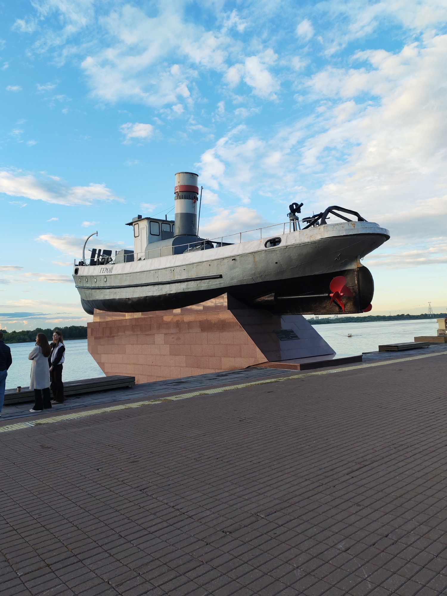 This media file captures a monument of a ship, likely a tugboat, on display outdoors. The ship, painted primarily in grey with a black hull, rests on a prominent reddish-brown stone pedestal. Visible on the side of the ship is the word "ГЕРОЙ" (Hero). The ship has a tall smokestack with red and white bands and a red propeller at its stern.
In the foreground, a paved walkway made of dark bricks leads towards the monument. To the left of the ship, three people are standing on the walkway, seemingly observing the monument. One person is wearing a dark jacket, and two women are wearing lighter-colored coats.
The scene is set against a backdrop of a wide body of water, possibly a river, with trees visible on the far bank. The sky above is a bright blue with scattered white clouds, suggesting it is daytime, likely late afternoon or early evening due to the quality of the light. The overall atmosphere appears calm and serene. The location is identified as Nizhny Novgorod, Russia.
