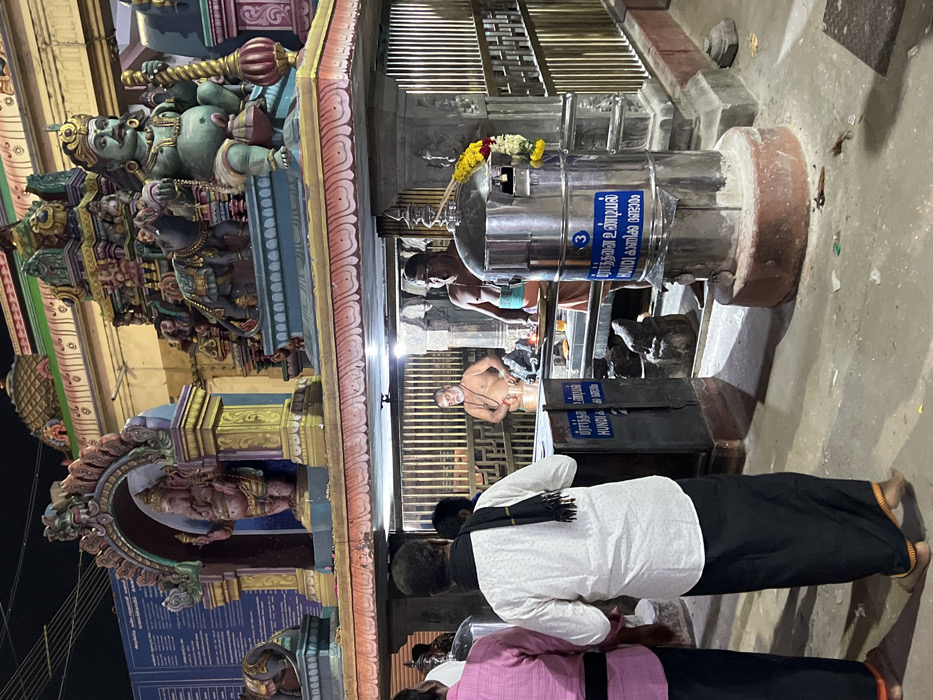 This image captures a nighttime or evening scene within a Hindu temple in Palani, India. In the foreground, two individuals, presumably devotees, are seen from behind, facing towards the temple's inner areas. One wears a white shirt with a black scarf and black lower garment, while the other wears a pink checkered shirt.
A prominent feature in the midground is a large, metallic cylindrical donation box, known as a *hundi*. It is marked with the number '3' and displays text in both Tamil and English. The Tamil text reads "நன்கொடை உண்டியல்" (Nankodai Undiyal), which translates to "Donation Hundi," and the English word "HUNDI" is also clearly visible.
Behind the hundi, two men, likely temple priests or attendants, are present. One is bare-chested and wears a sacred thread, facing towards the devotees. The other, partially obscured, wears an orange dhoti and appears to be performing duties or attending to offerings. Small statues or idols are placed at the base of the hundi.
The background showcases vibrant and intricate temple architecture, adorned with numerous colorful sculptures of deities and mythological figures, some resembling gopuram artistry. These figures are richly detailed, featuring multiple arms and traditional iconography. Artificial lighting illuminates these sculptures and the general area, casting a warm glow and highlighting the temple's craftsmanship against the darker surroundings. An information board is partially visible on the far left, though its text is illegible. The scene evokes a sense of devotion and ritual within a sacred space.