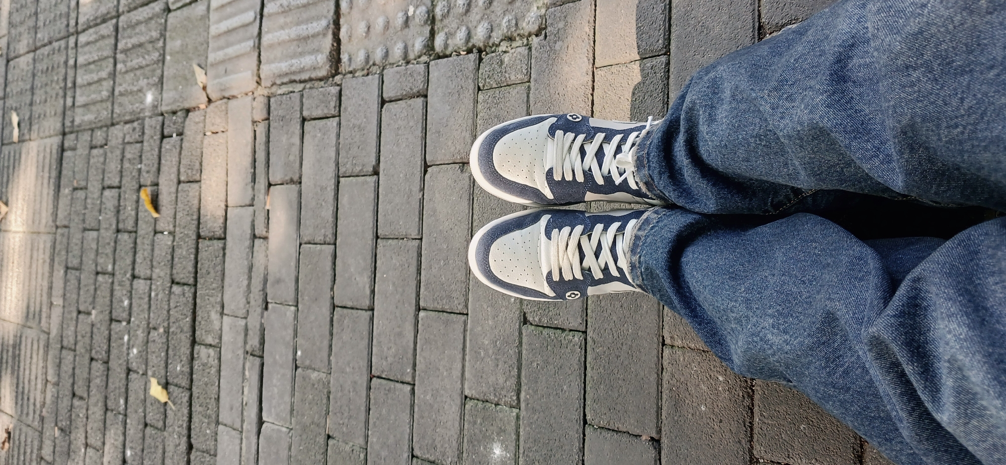 This image presents a top-down view of a person's feet and lower legs on a paved outdoor pathway in Yuzhong District, China. The person is wearing blue denim jeans, which appear to have a relaxed or loose fit, and two-toned sneakers.
The sneakers are primarily light cream or off-white, featuring perforated details on the toe box and side panels, contrasted with dark blue (likely navy) overlays and trim. They are secured with white laces. A distinct, stylized circular logo, resembling an '8' or infinity symbol, is visible on the outer side of each shoe, near the ankle area.
The ground is covered with a variety of gray paving stones, arranged in different rectangular patterns. Some sections of the pavement exhibit tactile paving, characterized by raised dots or lines, often used to aid visually impaired pedestrians. Scattered across the paving stones are several dry, yellowish-brown leaves, suggesting an autumn season.
Bright sunlight casts clear, angled shadows across the pavement and partially onto the shoes, indicating that the picture was taken during daytime, possibly morning or late afternoon. The scene captures a moment of standing or pausing in an urban pedestrian environment. No specific activity beyond standing is depicted, and no emotions are discernible.