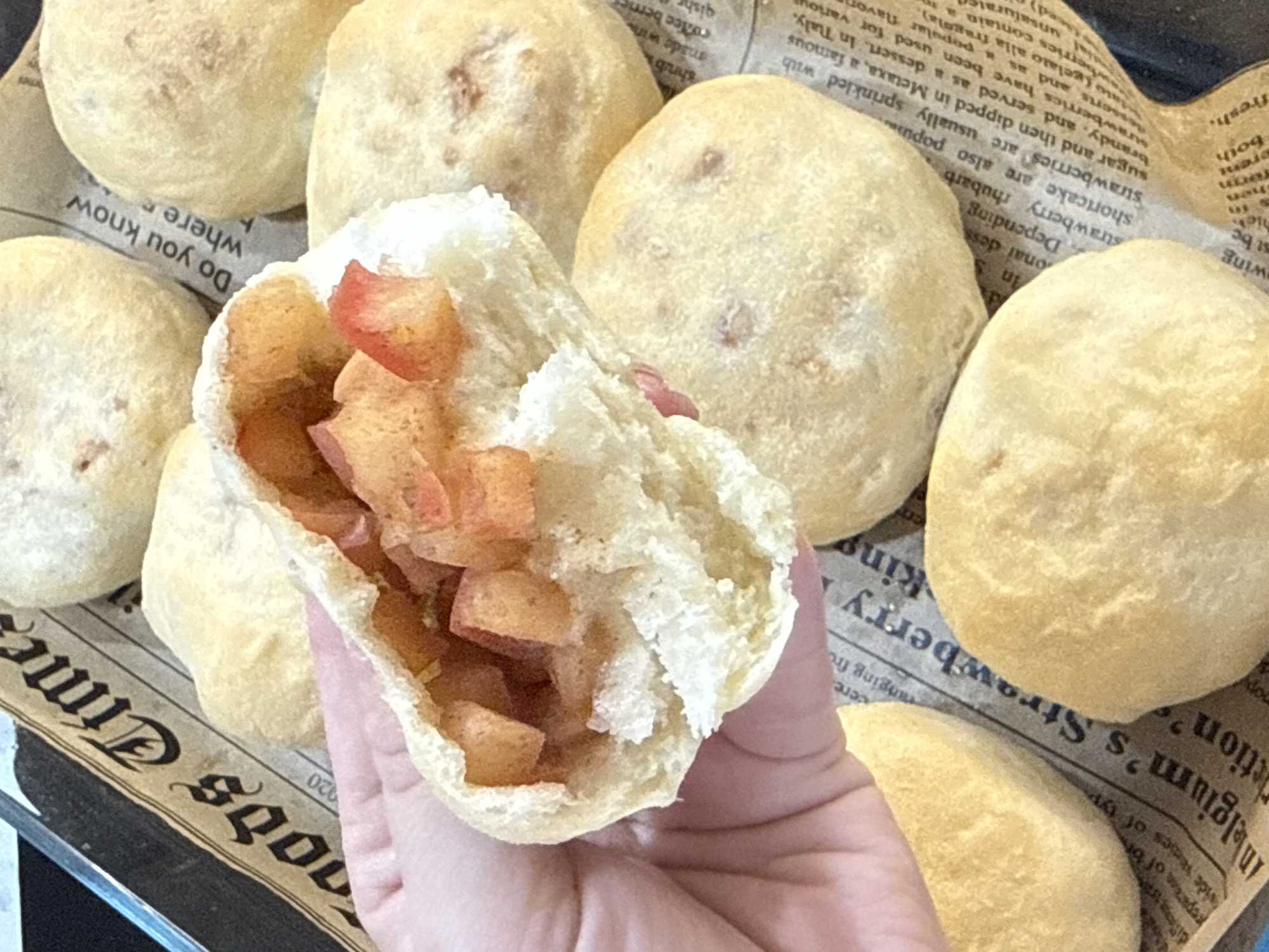 A close-up shot captures several round, light-colored pastries or buns, resting on a sheet of printed paper that resembles a newspaper. A human hand holds one of these pastries, torn open to reveal a filling of diced, reddish-orange pieces, likely cooked fruit such as peaches or apples. The interior of the pastry appears soft and white.
The other pastries are scattered around, displaying a uniform light golden-brown color and a slightly textured surface. The printed paper beneath them contains various articles and text fragments. Visible text includes phrases like "Do you know where," "Strawberry," "Belgium's," and mentions of "strawberry rhubarb shortcake," "strawberries are usually sprinkled with sugar and then dipped in Metaxa, a famous brandy, and served as a dessert. In Italy, strawberries have been used for various... (gelato alla fragola)... strawberries contain..." Other discernible words are "nods Time" (likely "Goods Times"), "920," "shrub," and "coffee berries."
The setting appears to be indoors, possibly a kitchen or dining environment, under natural or bright artificial light. No specific time of day, weather conditions, or emotional states are discernible. The image itself provides no visual cues to confirm the given location context of Lyubertsy, Russia.