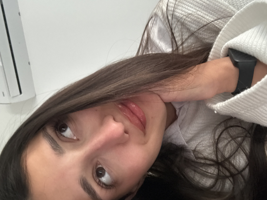 The image displays a close-up, angled indoor portrait of an adult female. She has long, dark brown hair, brown eyes, and is looking upwards and to the viewer's left with a subtle smile. Her chin is supported by the fingers of her right hand, which is partially visible. She wears a light-colored, textured long-sleeved garment, and a black digital smartwatch is present on her right wrist. The background consists of a plain white wall. A white wall-mounted air conditioning unit is visible in the upper right portion of the frame. The precise location within the city of Budva, Montenegro, cannot be determined from the visual information presented in the image.