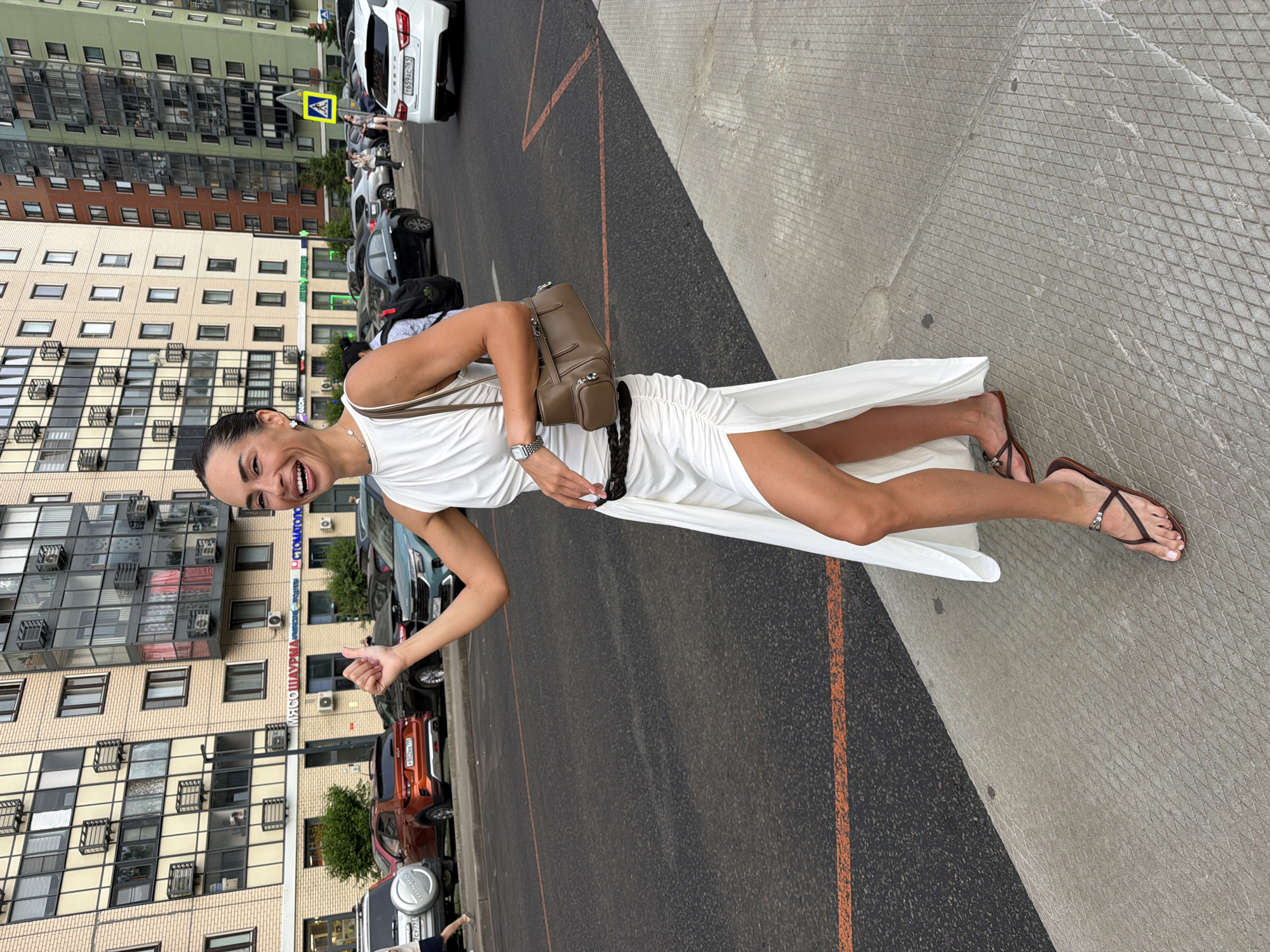 A woman in a white dress is standing on a sidewalk next to a street in an urban area. The woman is smiling and has her right arm raised, with her thumb up. She is wearing a white halter dress with a high slit on the left side, a brown purse slung over her shoulder, and brown flip-flops. Behind her, there are several cars parked along the street and a multistory building with many windows. The building appears to be residential or commercial, with visible signs. The sidewalk has a textured pattern. The street has orange markings. In the background, other people and vehicles are visible, indicating a public urban environment.