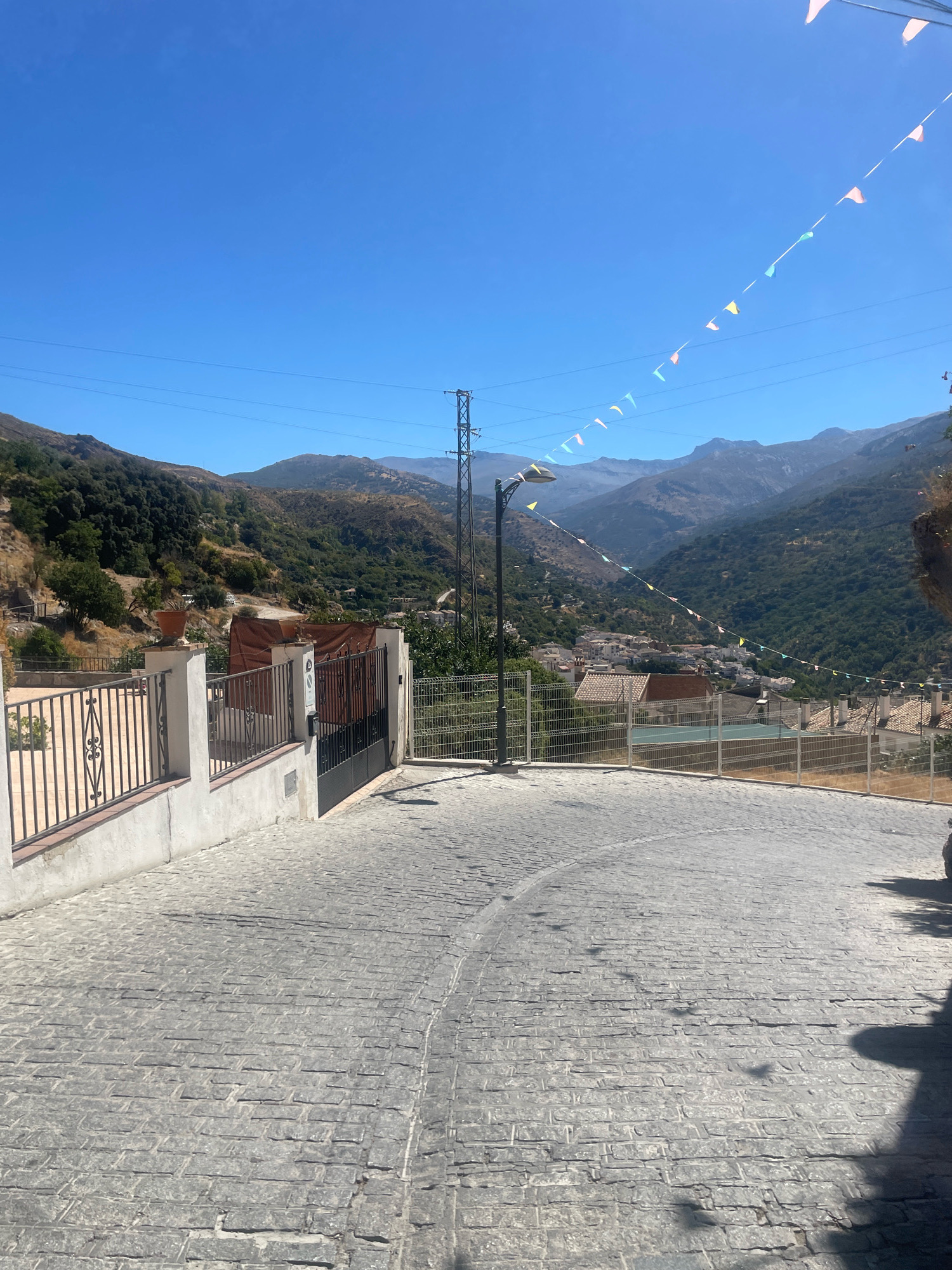 The image captures a sunlit daytime scene on a sloped, cobblestone street in Güéjar Sierra, Spain. The street surface is paved with rectangular gray stones arranged in a repeating pattern. On the left, a white wall with a black metal fence runs along the edge of the property, topped with ornamental posts and a potted plant. Beyond the fence, lush green hills dotted with trees and vegetation rise towards the sky. A tall metal electricity pylon stands prominently in the middle ground. Further in the distance, a cluster of white buildings with terracotta roofs forms a village nestled within a valley, surrounded by more verdant, mountainous terrain. Colorful triangular flags are strung across the sky, adding a festive touch to the scene. The sky is a clear, bright blue, and shadows from unseen objects are cast across the street, indicating the sun's position. The overall impression is one of a picturesque, rural Spanish village under a clear sky.