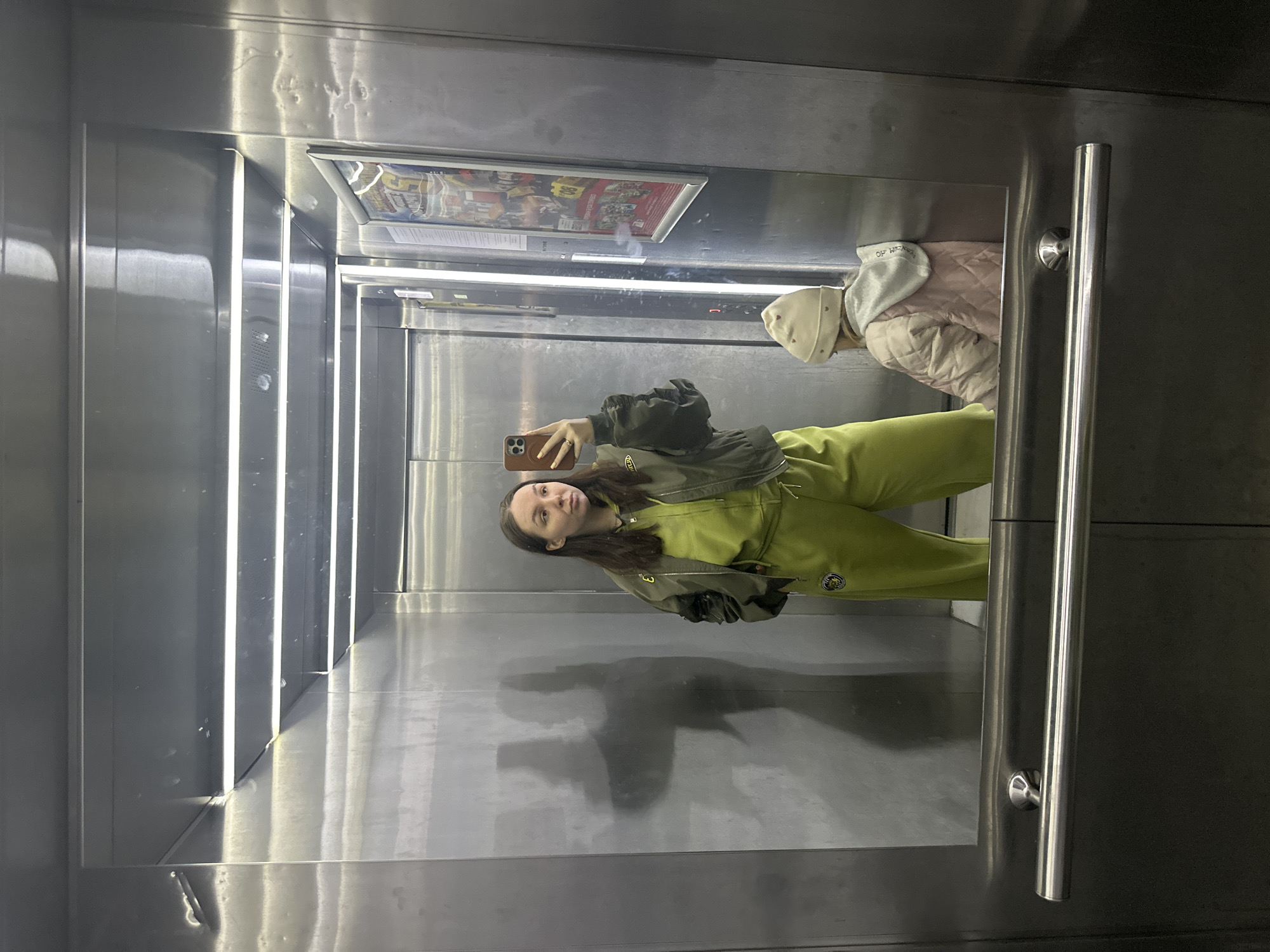 A young woman is taking a selfie in an elevator. She is wearing a green jumpsuit and holding a smartphone. The elevator has stainless steel walls and handrails. There's also a poster on the top part of the elevator. The location is unknown. The city of Chaikovsky, Russia is not clearly evident in the image.