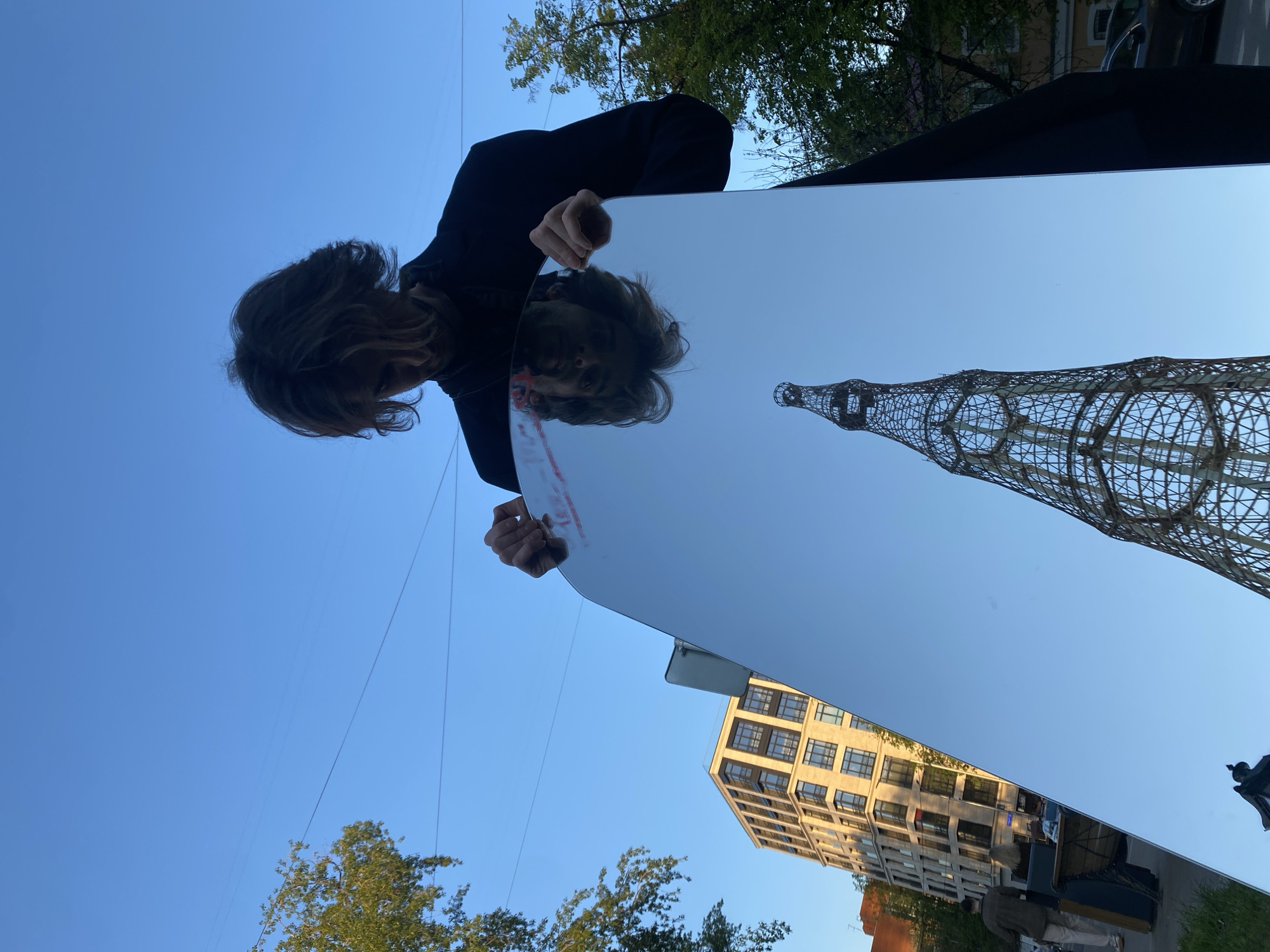 A low-angle shot reveals a person holding a reflective, rectangular mirror. The mirror reflects the Shukhov Tower in Moscow, Russia, alongside an apartment building. The person holding the mirror is viewed from above, with hair visible. The sky is clear blue.