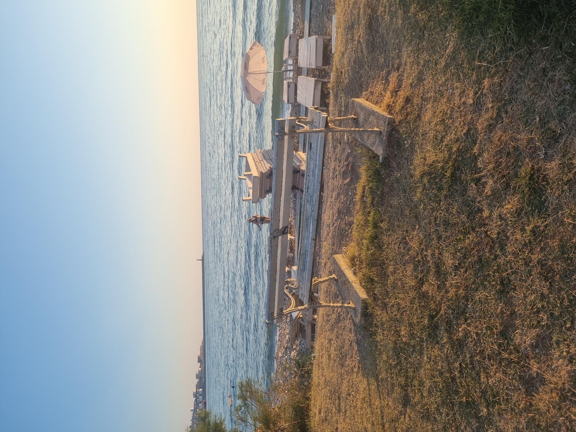 The image depicts a tranquil scene at a beach in Bar, Montenegro, during the late afternoon or early evening, as indicated by the warm, golden light casting long shadows. In the foreground, a weathered wooden bench with ornate metal legs sits on dry grass and soil. Behind the bench, a rocky beach stretches towards the sea. Several wooden lounge chairs and small tables are arranged near the water's edge, along with a light-colored umbrella providing shade.
A person in swimwear stands near the water on the right side of the frame. In the distance, the calm, blue sea meets a clear sky that transitions from a soft blue at the top to a pale yellow near the horizon. Along the coastline in the far distance, industrial structures with white cylindrical tanks and a lighthouse on a pier are visible, suggesting the presence of a port or industrial area. The overall atmosphere is one of peacefulness and relaxation, characteristic of a beachside setting at dusk.