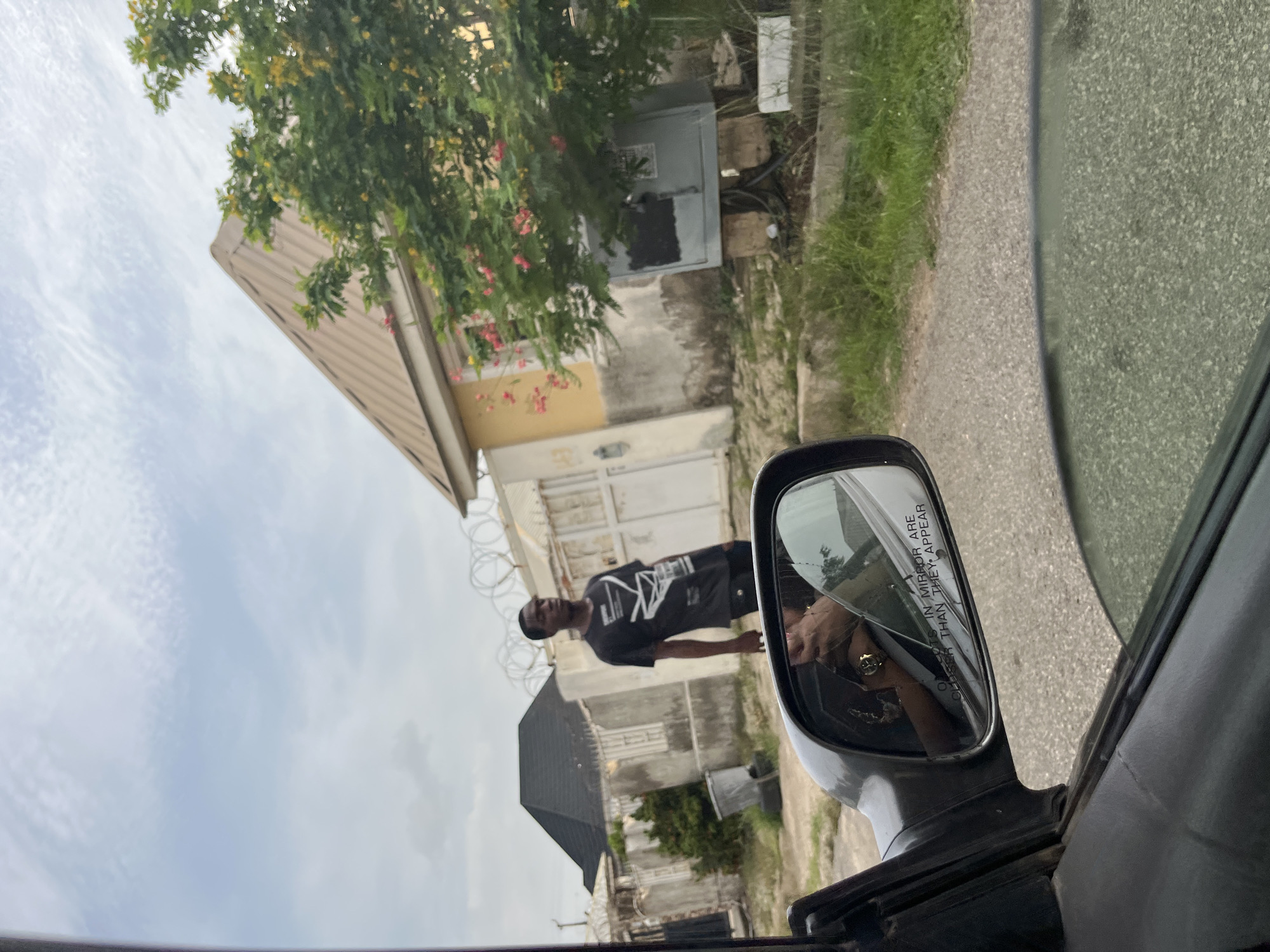 The image, taken from inside a vehicle, shows a residential street scene in what is believed to be Abuja, Nigeria. Through the side mirror, a young man in a black t-shirt is visible standing on the sidewalk. The reflection in the mirror also shows the person taking the photograph wearing a ring. Buildings are visible in the background, one with a beige exterior and a corrugated metal roof and another with a dark roof. Razor wire is secured on top of the fence that surrounds the buildings. The sky is overcast. The interior of the car is visible in the foreground.