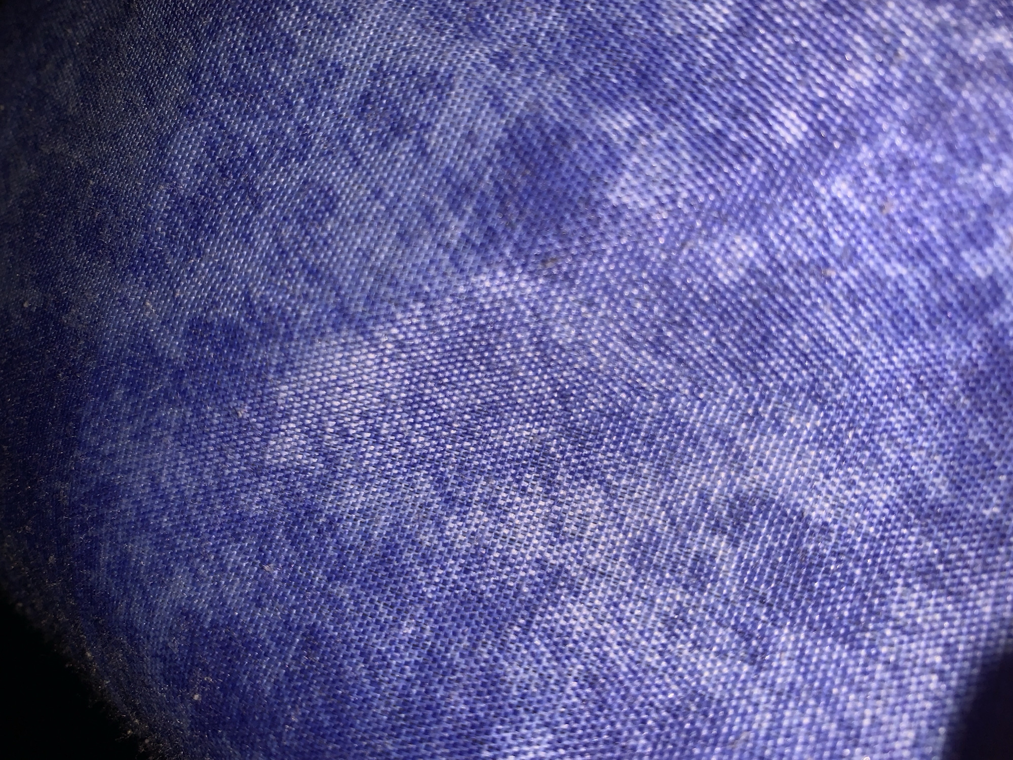 The media file displays a close-up, high-magnification view of a blue woven fabric. The primary subject is the textile's surface. No people or other distinct objects are visible, focusing solely on the material's texture.
No specific activity or event is taking place; the image is a static representation of the fabric.
Notable details include the fabric's color, which is predominantly blue but varies significantly across the surface. There are darker, richer blue sections and lighter, faded areas that sometimes show a purplish tint. This variation in color suggests either natural wear and fading, a stone-washed finish, or uneven illumination. The fabric exhibits a fine, intricate woven pattern, appearing as a grid of interlocked threads, typical of denim or similar durable textiles. Small, dark specks, possibly dust or lint, are sparsely distributed across the fabric surface. The lighting appears somewhat uneven, casting subtle shadows and highlights that emphasize the textile's woven structure. There are no visible location cues within the image itself, nor any discernible time of day, weather conditions, or emotional context. No text is visible in the media file.