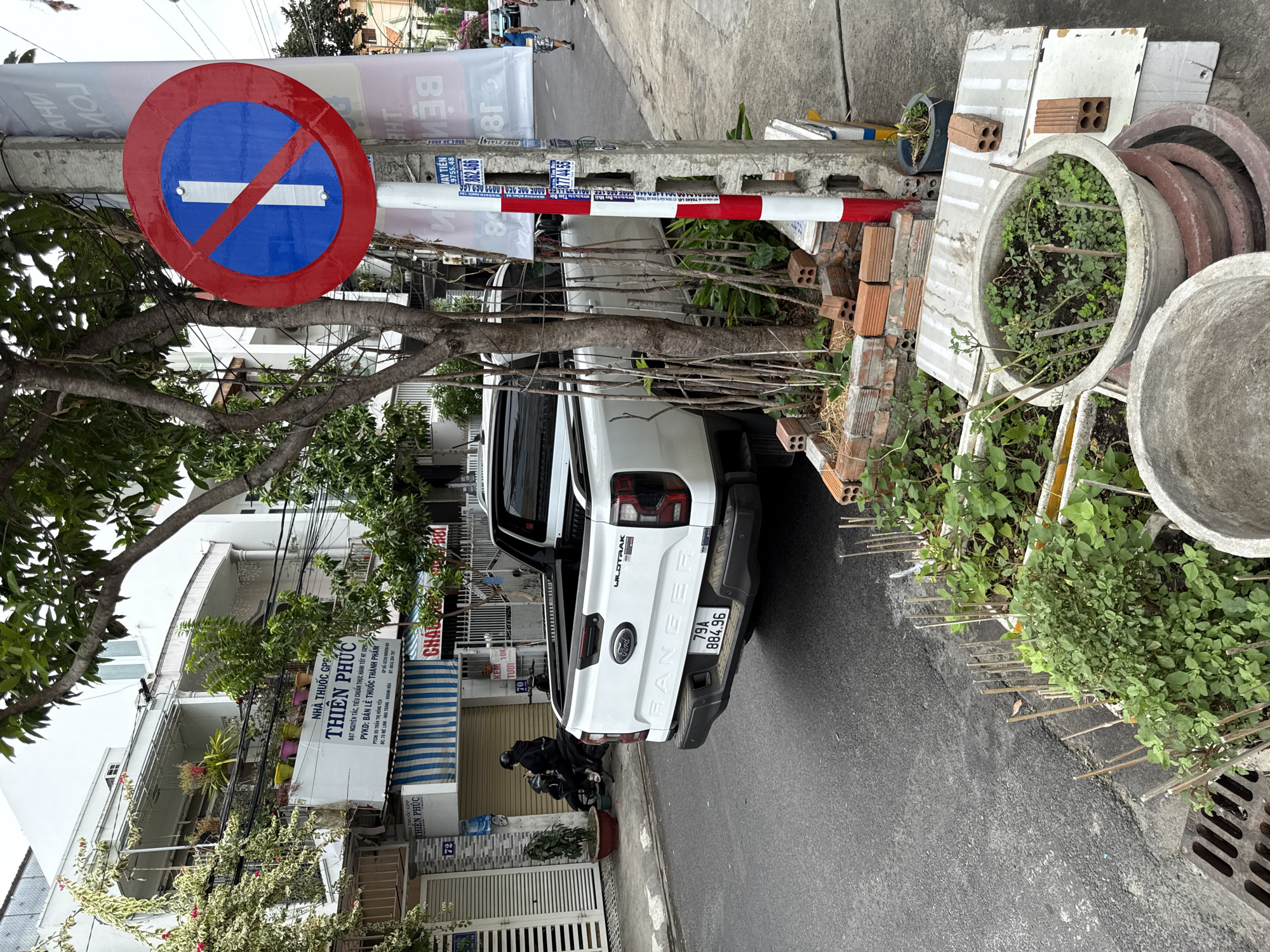 This image shows a street scene in Nha Trang, Vietnam. In the foreground, a white Ford Ranger pickup truck is parked on the right side of the road. The truck has a license plate that reads "79A 884.96". To the left of the truck, a sidewalk is visible with several potted plants. Further down the street, a person on a motorcycle is seen. On the right side of the image, a traffic sign indicating "no stopping" is mounted on a pole. Behind the truck, buildings line the street. The weather appears to be overcast.