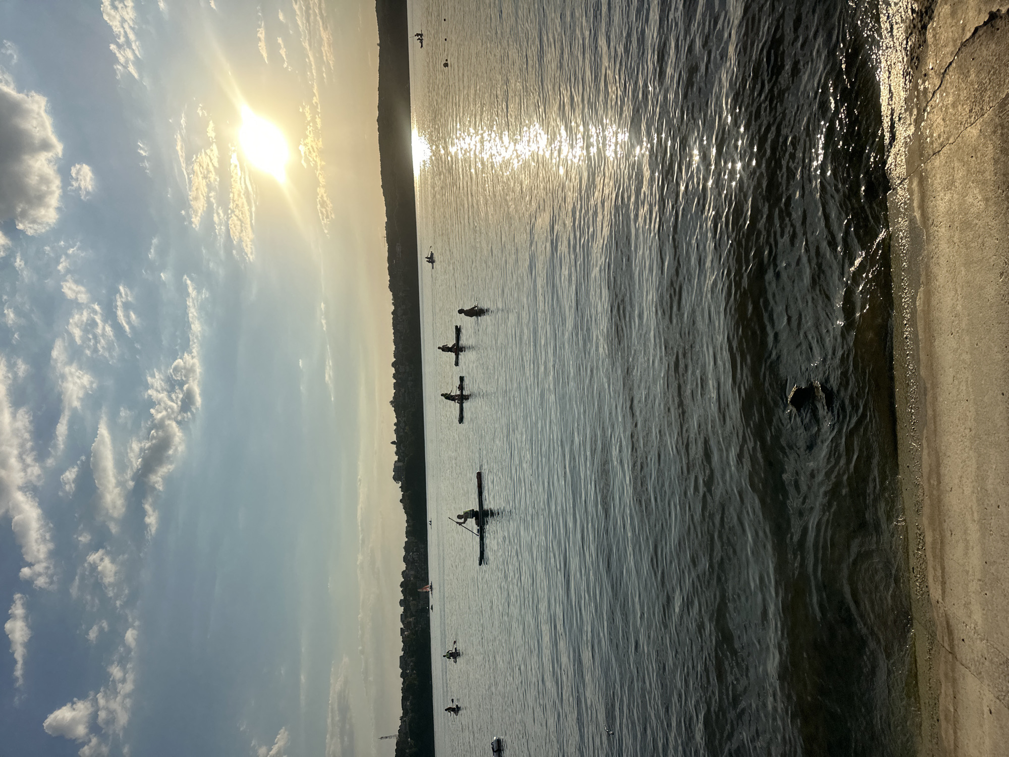 The image captures a tranquil scene on Ternopil Pond in Ternopil, Ukraine, during what appears to be a late afternoon or early evening. The sun is setting, casting a warm glow across the water and creating a shimmering reflection. Several people are engaged in water activities, primarily on paddleboards.
In the foreground, a concrete embankment or shoreline is visible, with the dark water lapping gently at its edge. The water itself is calm, with subtle ripples and reflections of the sky.
Further out on the pond, multiple individuals are dispersed. The most prominent is a person on a paddleboard in the center of the frame, paddling forward with a single-bladed oar. To their left, two other paddleboarders are also visible, and to the right, another paddleboarder is present. In the distance, more individuals can be seen on paddleboards, as well as a few sailboats or windsurfing sails adding to the serene atmosphere.
The background features a treeline along the far shore, dotted with houses and buildings, indicating a populated area bordering the pond. The sky is a mix of blue with scattered white and gray clouds, illuminated by the setting sun which is partially obscured by clouds but still quite bright, creating a soft lens flare effect. The overall impression is one of peaceful recreation and natural beauty in an urban setting.