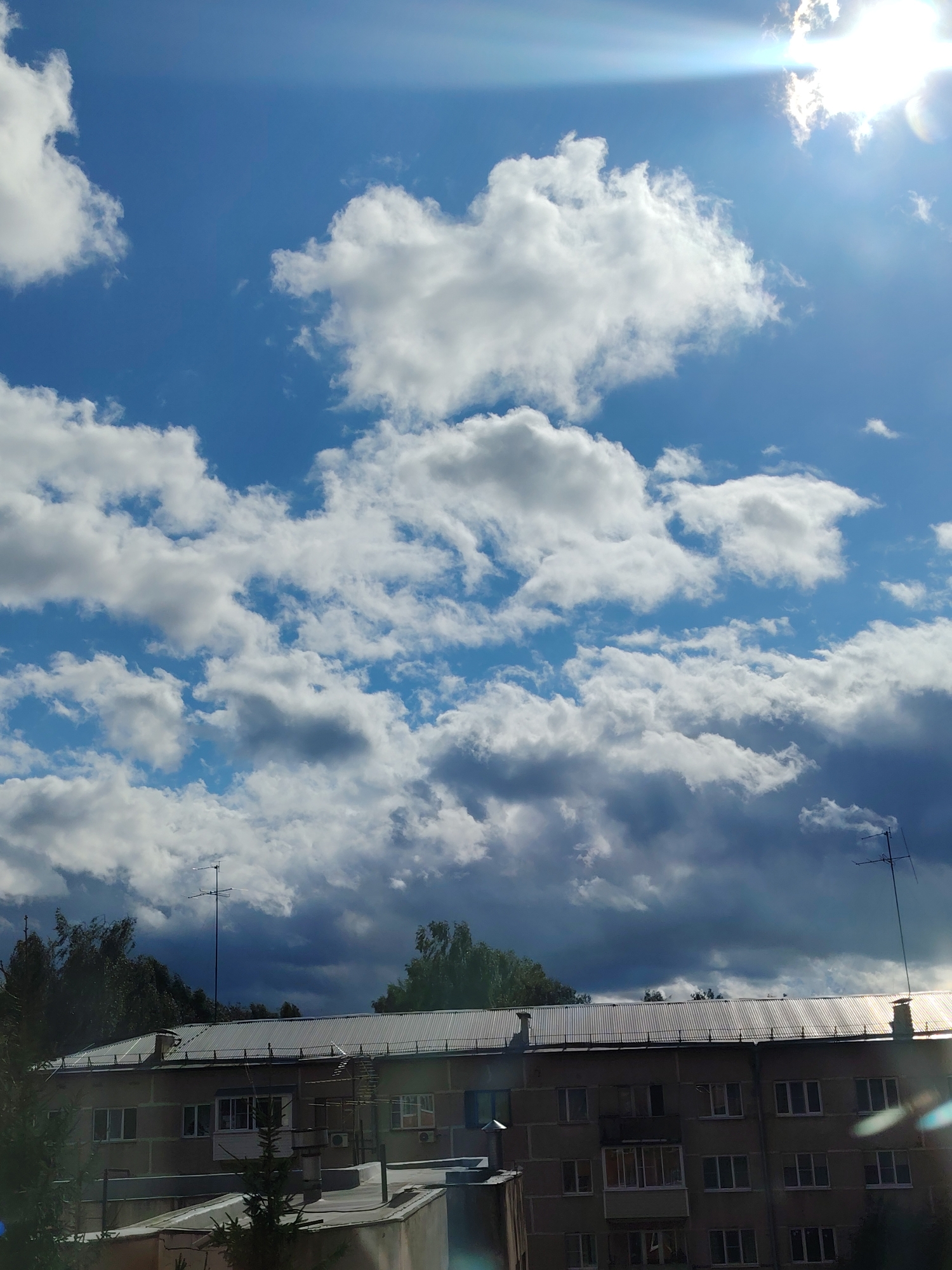 The image depicts a residential building with a corrugated metal roof under a dynamic sky filled with cumulus and dark stratus clouds. The sun is visible in the upper right quadrant, casting a lens flare across the sky. Two television antennas are mounted on the roof. Trees frame the building on the left and right sides. The scene appears to be an exterior view of an apartment building in Konakovo, Russia, with signs of urban infrastructure and vegetation.