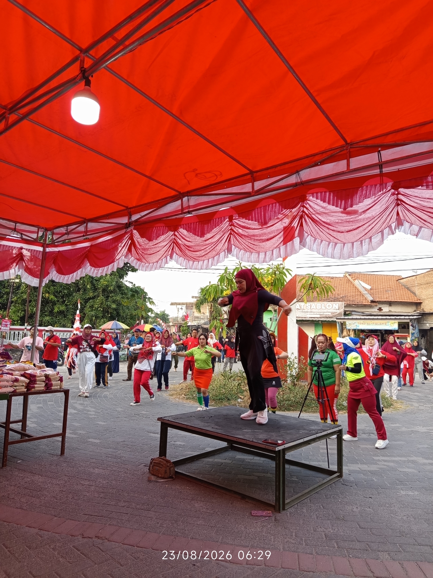 On August 23, 2026, at 06:29 AM, a group of people are participating in an outdoor aerobic activity under a large red tent in Canggu, Indonesia. The scene is set on a paved area, possibly a street or a public square, with buildings and trees visible in the background.
The focal point of the image is a woman in a dark hijab and black athletic wear, who is leading the aerobics class from a raised platform. She is in mid-movement, with her right arm extended and her body slightly bent. Another woman in a bright green shirt and a red skirt is positioned next to her on the platform, operating a video camera on a tripod, likely recording the event. A third woman in a red and yellow sports outfit stands to the right of the leader, also participating in the exercise.
Behind the platform, a crowd of about 15-20 individuals, mostly women, are following the aerobics routine. They are dressed in various activewear, many in red, white, and green, suggesting a coordinated event or a national theme. Some are wearing hijabs, and others are not. Several are holding small flags or wearing accessories with Indonesian colors.
To the left of the frame, there are tables displaying stacks of what appear to be sacks of rice or grains. Further in the background, a street lined with buildings and dense green trees can be seen. The overall atmosphere suggests a community event or a morning fitness gathering. The red tent provides shade and shelter for the participants, with red and white decorative fabric draped from its edges.