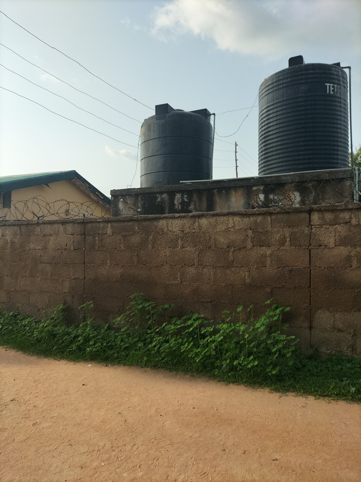 The image depicts an outdoor scene, likely in Jalingo, Nigeria. A rough-textured, brown brick wall dominates the foreground and mid-ground, with a dirt path and some green vegetation running along its base. Behind the wall, two large, dark water tanks are visible on a concrete platform. The tank on the right has the word "TET" visible on it. Barbed wire is strung across the top of the wall. A portion of a yellow building with a green roof is visible to the left, behind the wall. The sky above is a pale blue with scattered white clouds, and several thin wires are strung across the sky. There are no people or animals visible in the image.
