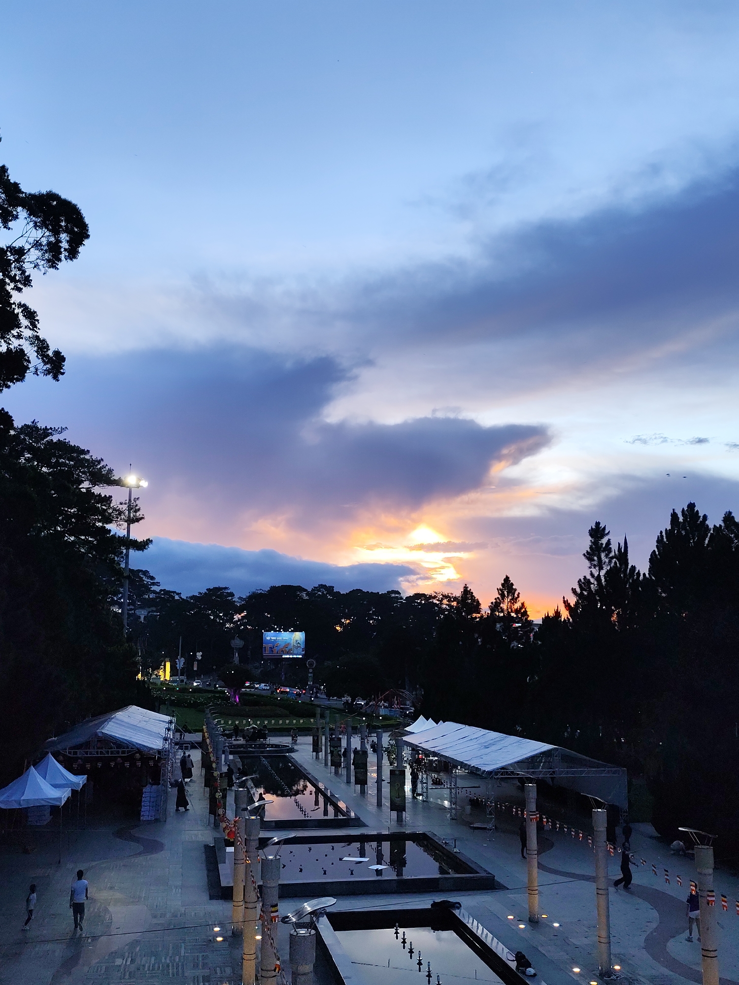 The image depicts a view of the Da Lat city park at dusk in Da Lat, Vietnam. The sky features a gradient of blue, purple, and orange hues. In the middle ground, there is a tree line with silhouettes of trees against the horizon.
In the foreground, a paved pedestrian area with multiple rectangular water features is visible. Vertical pillars, some adorned with flags or banners, line the walkway. Several temporary structures, possibly tents or canopies, are set up along the sides of the paved area. Some pedestrians are walking within the park.
In the background behind the tree line, a large electronic billboard is lit up. Traffic and other city lights are visible in the distance.